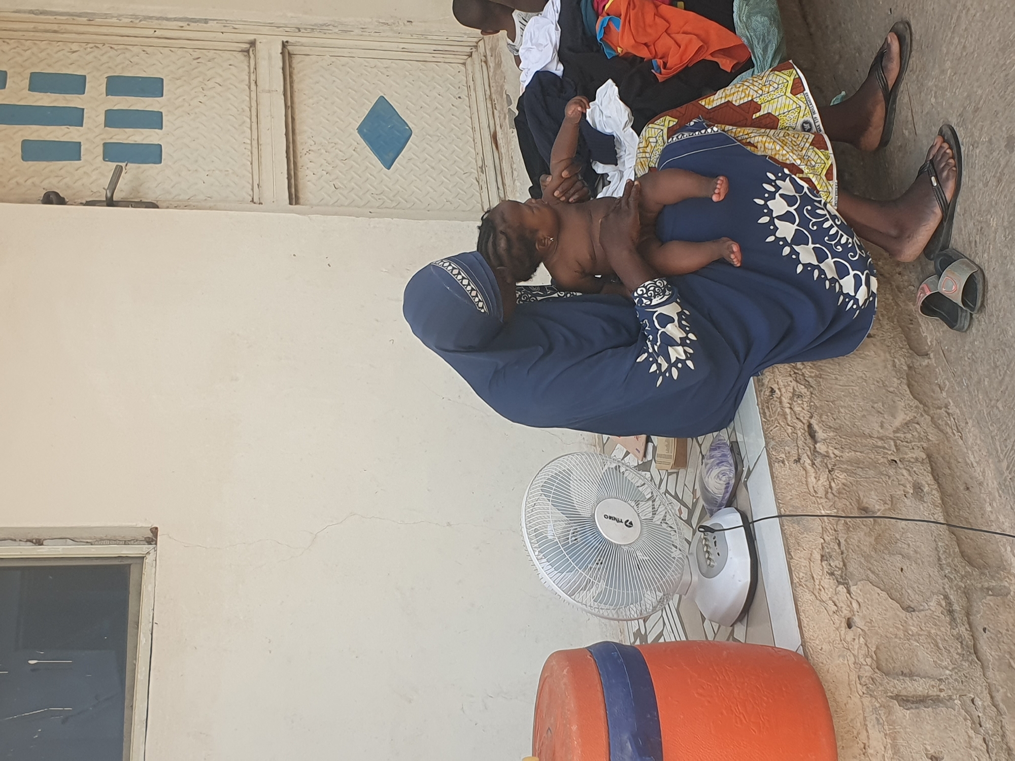 The image portrays a domestic scene in Lokoja, Nigeria, under bright daylight, likely outdoors or in a semi-enclosed space. A woman, wearing a dark blue, long-sleeved garment with white patterned sleeves and a matching head covering, is seated on the ground. She gently holds a small, shirtless baby with dark hair on her lap, suggesting a moment of care or feeding. Her feet, adorned with dark flip-flops, are visible.
In the background, a white metal door or gate with light blue rectangular and diamond designs is visible, featuring a metal latch. Nearby, other individuals are partially seen, indicated by legs and colorful clothing, suggesting more people resting in the shaded area. The ground transitions from a smoother, possibly tiled or concrete surface in the foreground to a rougher, earthy texture further back.
To the right of the woman, a white oscillating fan is positioned on the ground, with the brand "OPTIMO" visible on its grille, implying warm weather conditions. An orange barrel with a blue band is partially visible in the lower right corner, alongside a white power strip or similar electrical device. The light-colored walls and the overall setting suggest a practical, everyday environment. The scene evokes a calm, nurturing atmosphere.