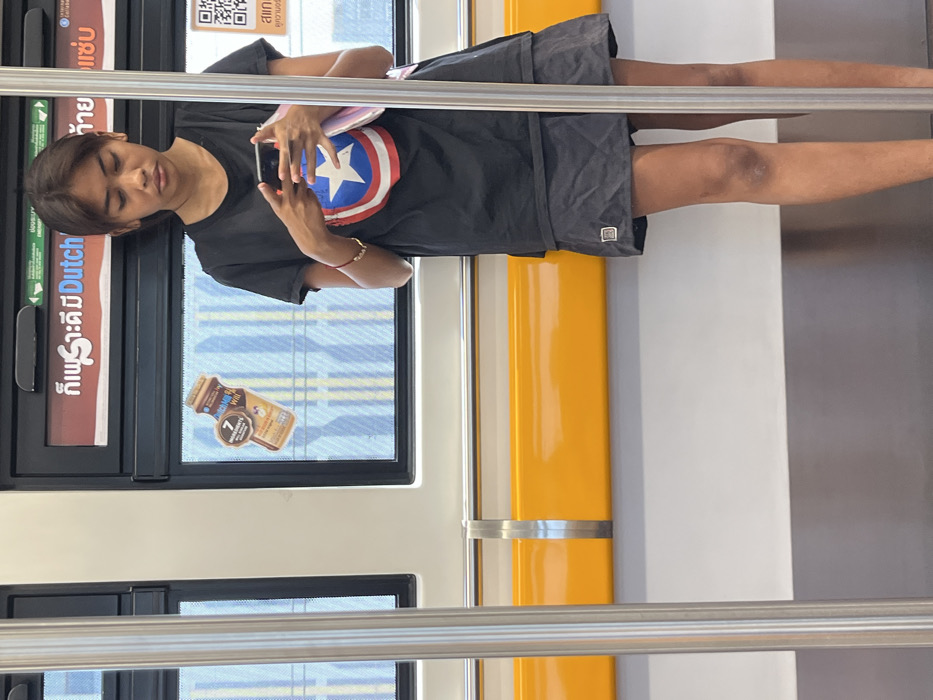 An image taken inside a public transit train, likely the BTS Skytrain, in Samut Prakan, Thailand, depicts a young female subject. The subject has dark hair and a light brown complexion. She is wearing a black short-sleeved t-shirt with a prominent Captain America shield logo on the chest, a dark grey or black pleated skirt, and a gold-colored bracelet on her right wrist. She is standing upright, holding a dark-colored smartphone in both hands and looking down at its screen. A light-colored object, resembling a notebook or small bag, is held in her left arm.
The train interior features white walls, a continuous horizontal yellow handrail, and a vertical silver support pole in the foreground. Through the train windows, a blurred exterior with vertical blue and yellow striped patterns is visible, suggesting movement. Advertisements are affixed to the windows; one at the top is brown-toned with Thai text including "Dutch Mill," and another lower down features a brown bottle graphic for "Dutch Mill 7 Ingredients Will." A partial QR code is visible to the far right of the frame. The lighting within the train appears bright and even.