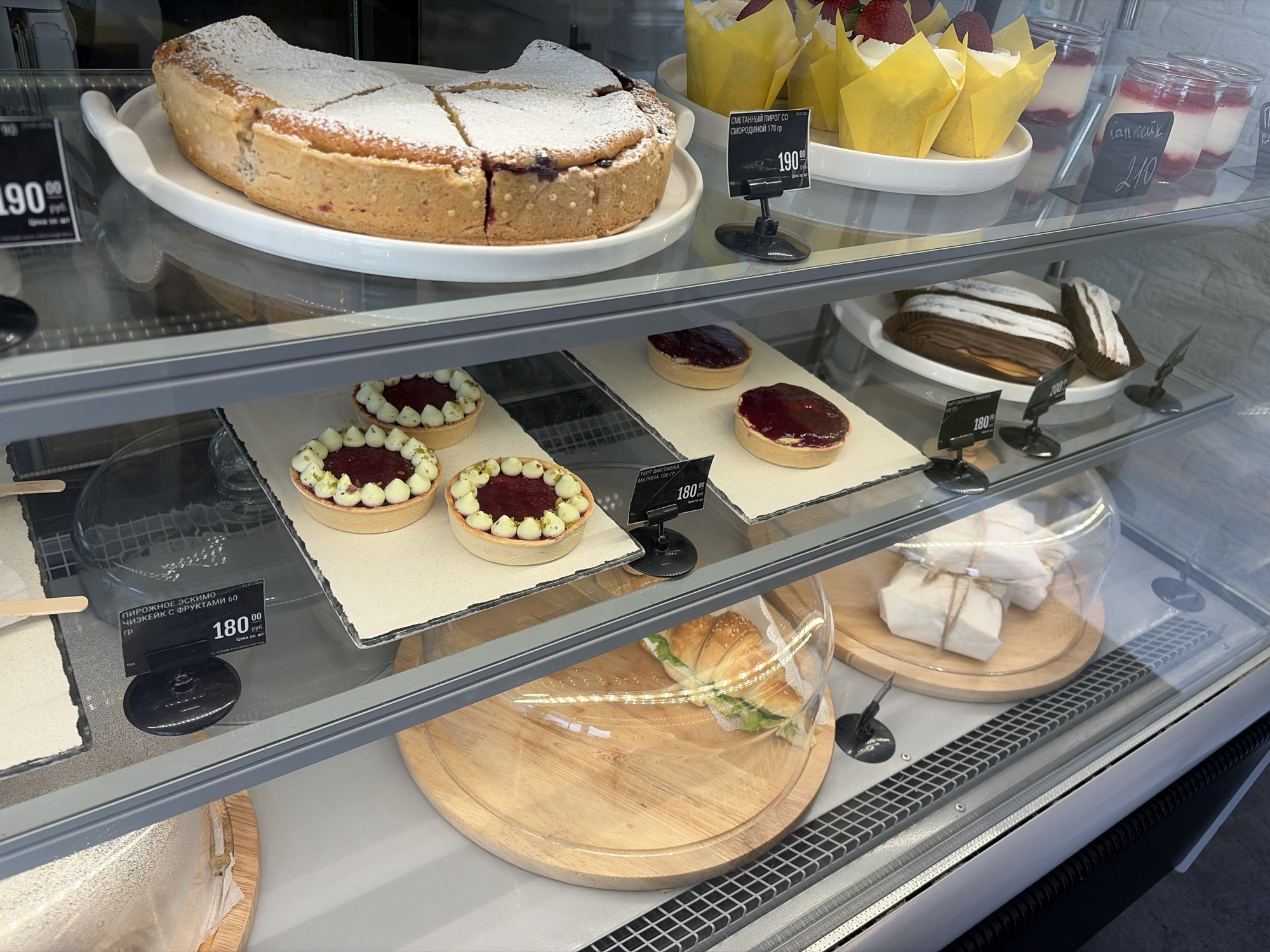 The image shows a refrigerated display case filled with various desserts and pastries. The case appears to be in a shop or bakery in Novopokrovskaya, Russia, based on the Cyrillic text on the price tags. The top shelf features a large pie dusted with powdered sugar, strawberry and raspberry cupcakes in yellow wrappers, and small jars of what appear to be panna cotta with red fruit sauce. The second shelf has small tarts with red filling and white decorations, as well as éclairs. The bottom shelf contains ice cream popsicles, and a sandwich. Price tags are visible for most of the items. The environment is clean and well-lit.