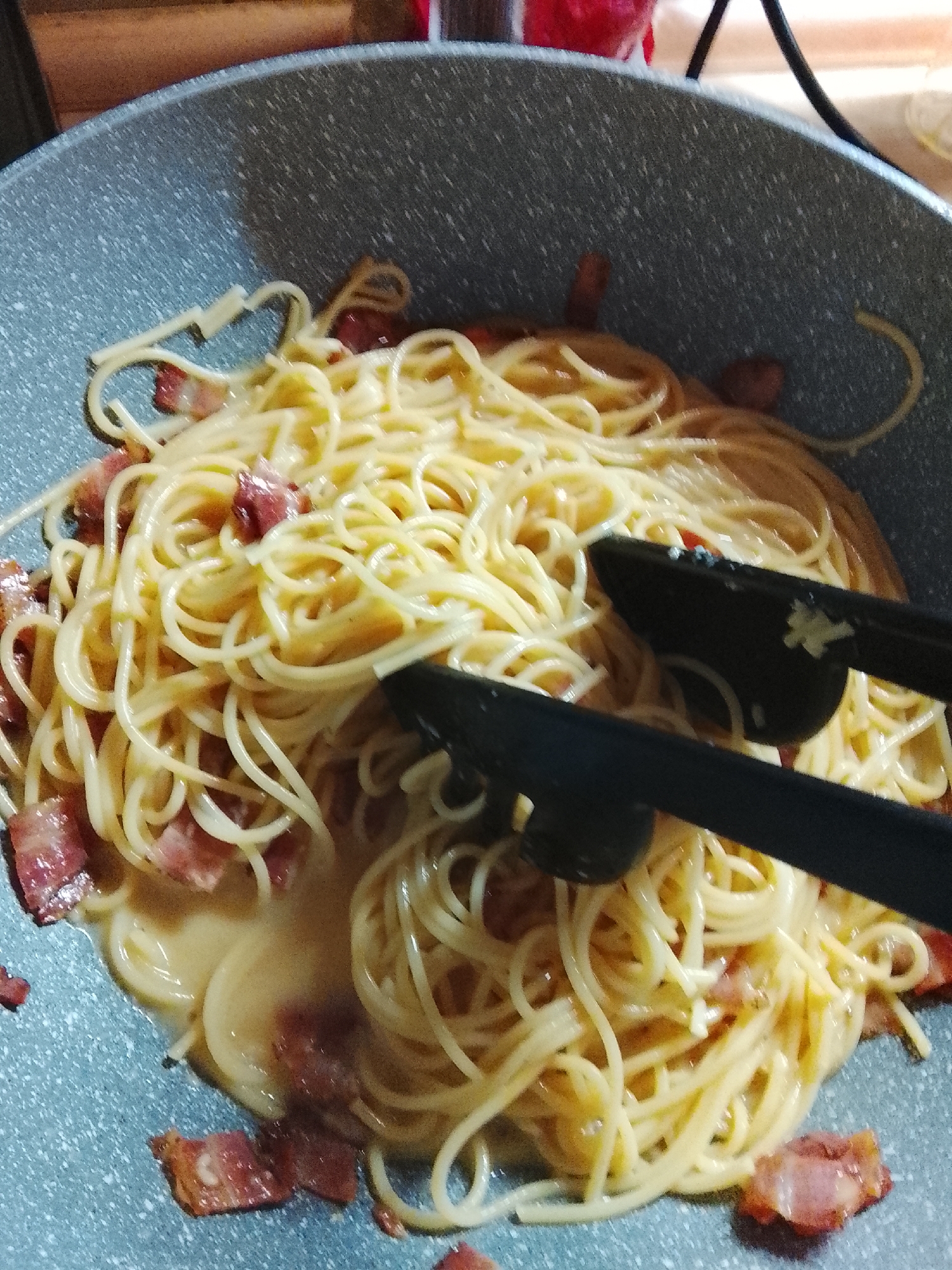 A close-up, overhead shot shows a large quantity of spaghetti with bacon and sauce in a speckled gray pan. The pasta is golden yellow and coiled in a messy pile. Small pieces of cooked bacon, red and white, are mixed throughout the spaghetti. A creamy, light yellow sauce coats the pasta and pools at the bottom of the pan. Black tongs are positioned on the right side of the pan, holding a portion of the spaghetti. The pan has a speckled gray interior. The background is blurred and out of focus, showing a wooden surface and a black cord. The image is taken from a high angle, looking directly down into the pan. There are no people visible in the image.