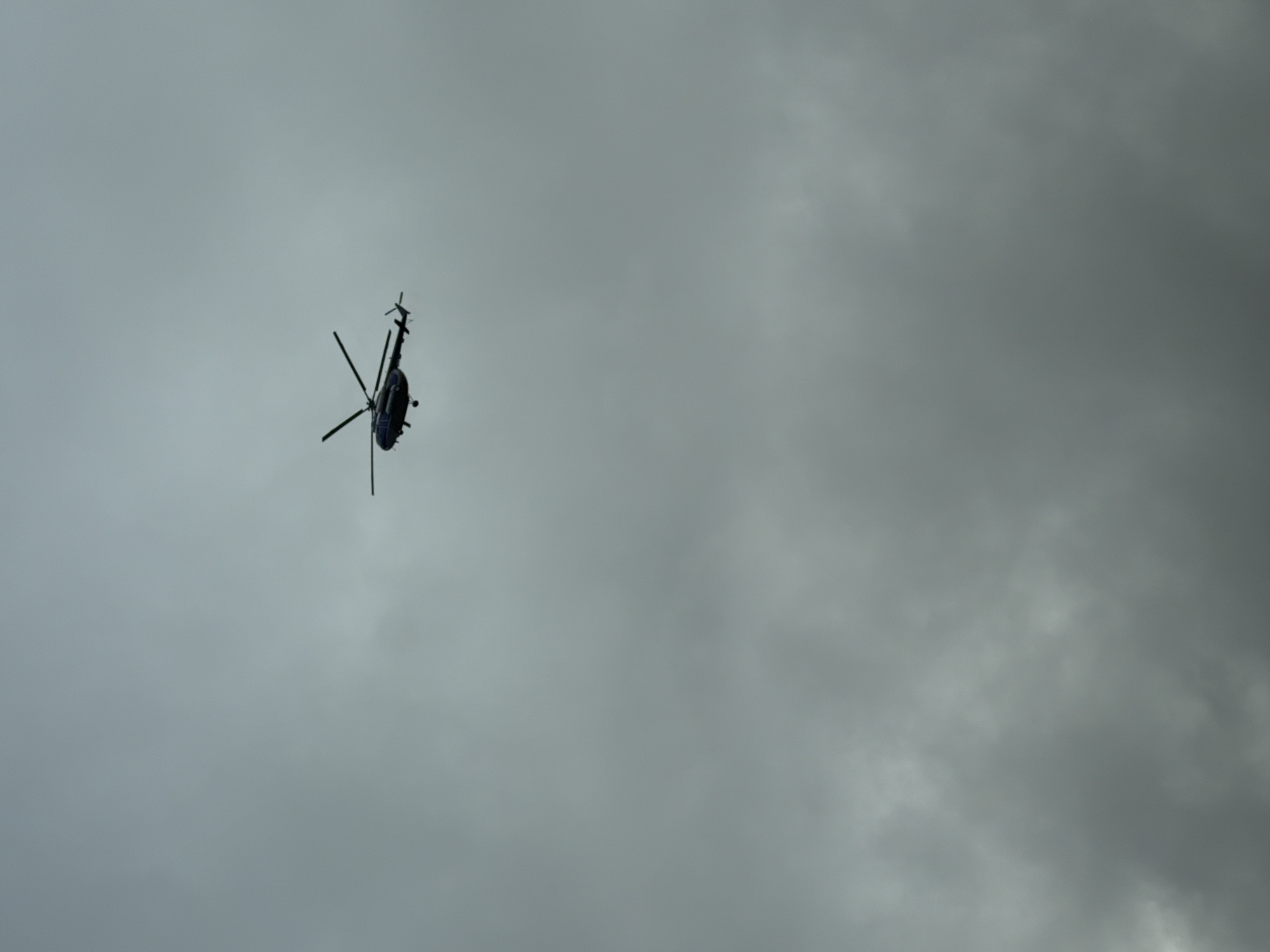 A helicopter is silhouetted against a gray, overcast sky. The location is Chaikovsky, Russia.