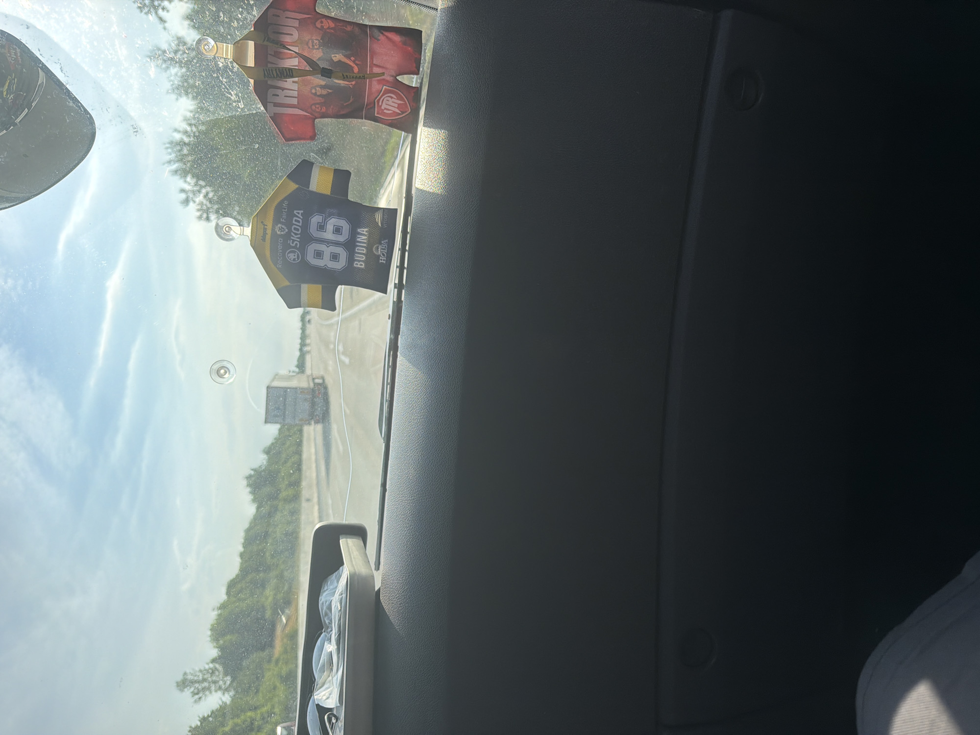 The image shows a view from inside a vehicle, looking out through the windshield. The vehicle appears to be traveling on a highway. In the foreground, on the dashboard, are two novelty air fresheners shaped like sports jerseys. One is black and yellow with the number 86 and the word "ŠKODA" and "BUDINA" visible. The other is red with "TRAKTOR" written on it and an image of several people.
Outside the vehicle, the highway stretches into the distance, with trees and greenery lining the sides. Several trucks are visible on the highway, including one directly ahead in the middle lane. The sky is blue with some scattered clouds. There is a circular suction cup mark on the windshield.
The provided context suggests the location is Hranice, Czechia. Based on this, the scene depicts a typical highway travel experience in that region. No people are clearly identifiable within the vehicle or as pedestrians. The focus is on the interior dashboard elements and the road ahead.