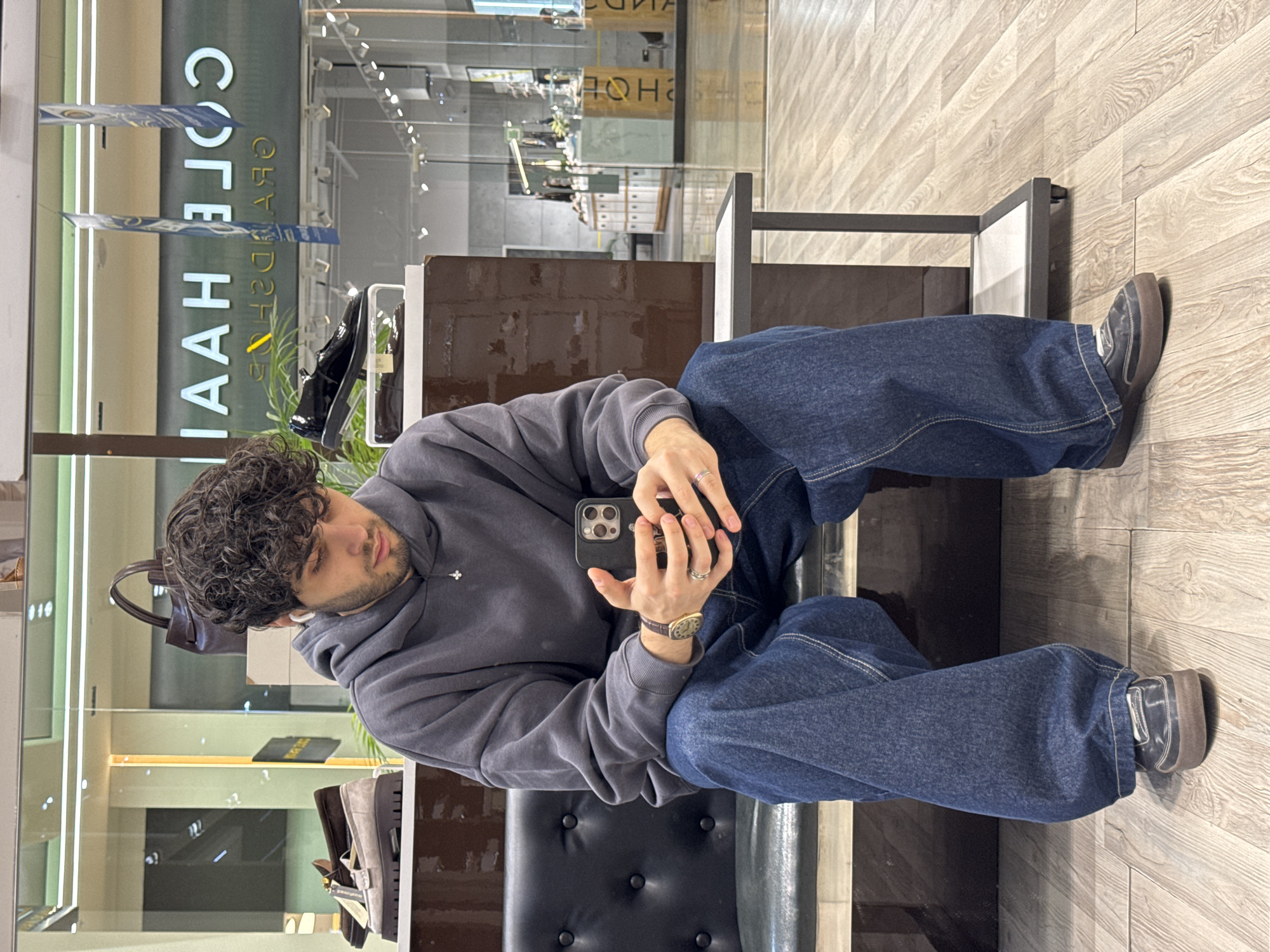 A young man is captured sitting on a black, tufted bench within a retail store. He has dark, curly hair and a short beard, and is dressed in a dark grey hooded sweatshirt adorned with a small white emblem, paired with baggy blue denim jeans. On his left wrist, he wears a gold-toned watch with a dark strap, and he has rings on his fingers. He is focused on a black smartphone held in both hands.
The setting features light-colored wooden floorboards and bright indoor lighting. Behind the man, a large glass partition or storefront displays the reversed text "COLE HAAN" and "SHOES," identifying the store as likely a Cole Haan establishment. The background reveals various shoes and handbags displayed on shelves and pedestals, consistent with a fashion or shoe boutique. The man appears calm and relaxed, engaged with his phone, possibly taking a break from shopping or waiting for someone. The time of day is not explicitly indicated, but the interior is brightly lit.