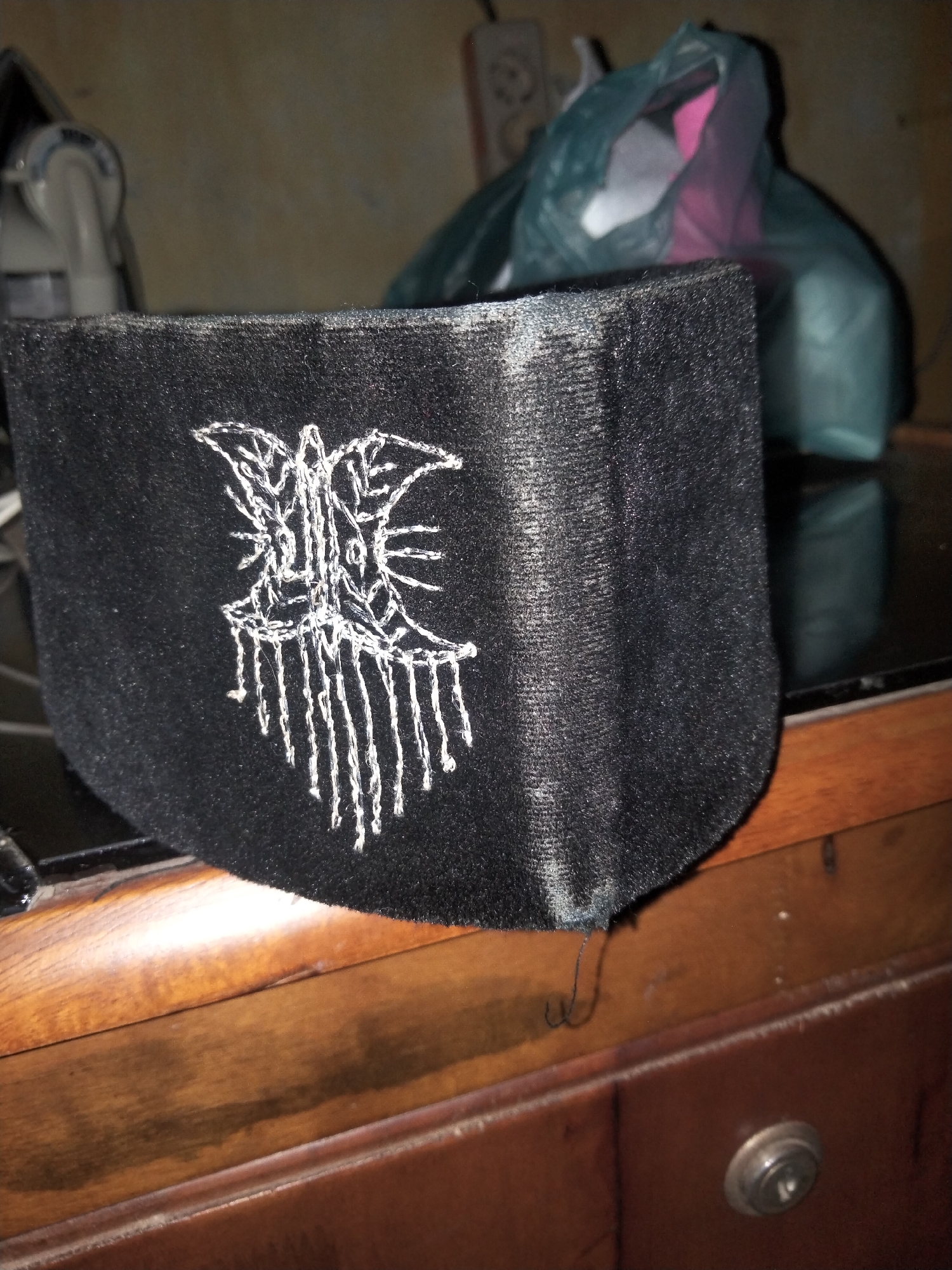 The image shows a close-up view of a black, textured fabric object with a white embroidered design. The design resembles a stylized bat or moth with intricate lines and dangling elements at the bottom. The fabric appears to be a pouch or cover with a curved bottom edge. The object is placed on a dark, reflective surface, likely a table or countertop, which sits on a wooden dresser. In the background, a blurred teal plastic bag is visible, along with what appears to be an appliance or electronic device with a white housing on the left side. The environment appears to be an indoor setting with dim lighting, suggesting a residential or workshop area in Payagaboh, Indonesia.