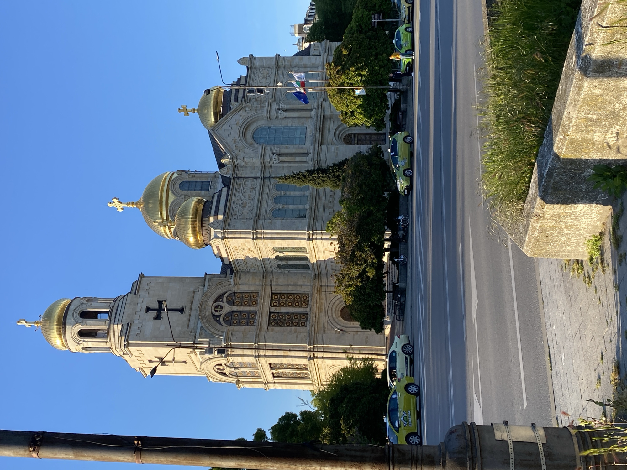 The image captures the Cathedral of the Dormition of the Mother of God in Varna, Bulgaria, from an elevated perspective, likely a bridge or raised walkway. The cathedral, a large, ornate structure with golden domes topped with Orthodox crosses, dominates the left side of the frame. A street runs alongside the cathedral, lined with green taxi vehicles. Flags fly near the entrance of the cathedral. The sky is clear and blue. A concrete barrier and some metal pipes are visible at the bottom of the image, suggesting the viewpoint's infrastructure. Vegetation, like grass, grows on the side of the street. There are no visible people in the frame.