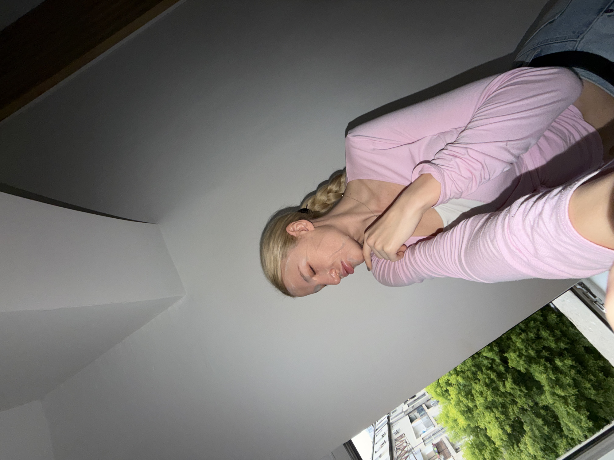 A woman with blonde hair in a braid is seen from a low angle. She wears a pink long-sleeved shirt and jeans. She appears to be looking down or has her eyes closed. Outside the window, there are trees and buildings that could be in an urban area. The location is Belgrade, Serbia.