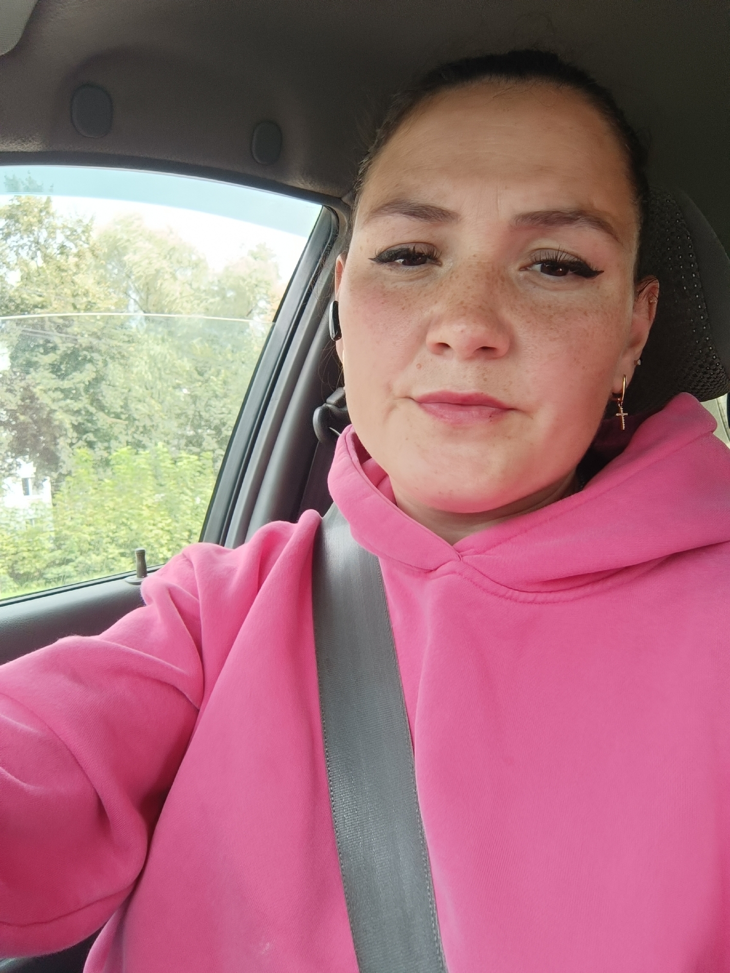The image is a selfie taken by a woman inside a car. She is wearing a pink hoodie and a seatbelt is visible across her chest. The woman has freckles and is wearing earrings. Outside the car window, trees and a building can be seen, suggesting an outdoor setting within a city. The scene appears to be in the city of Chaikovsky, Russia, based on the provided context.