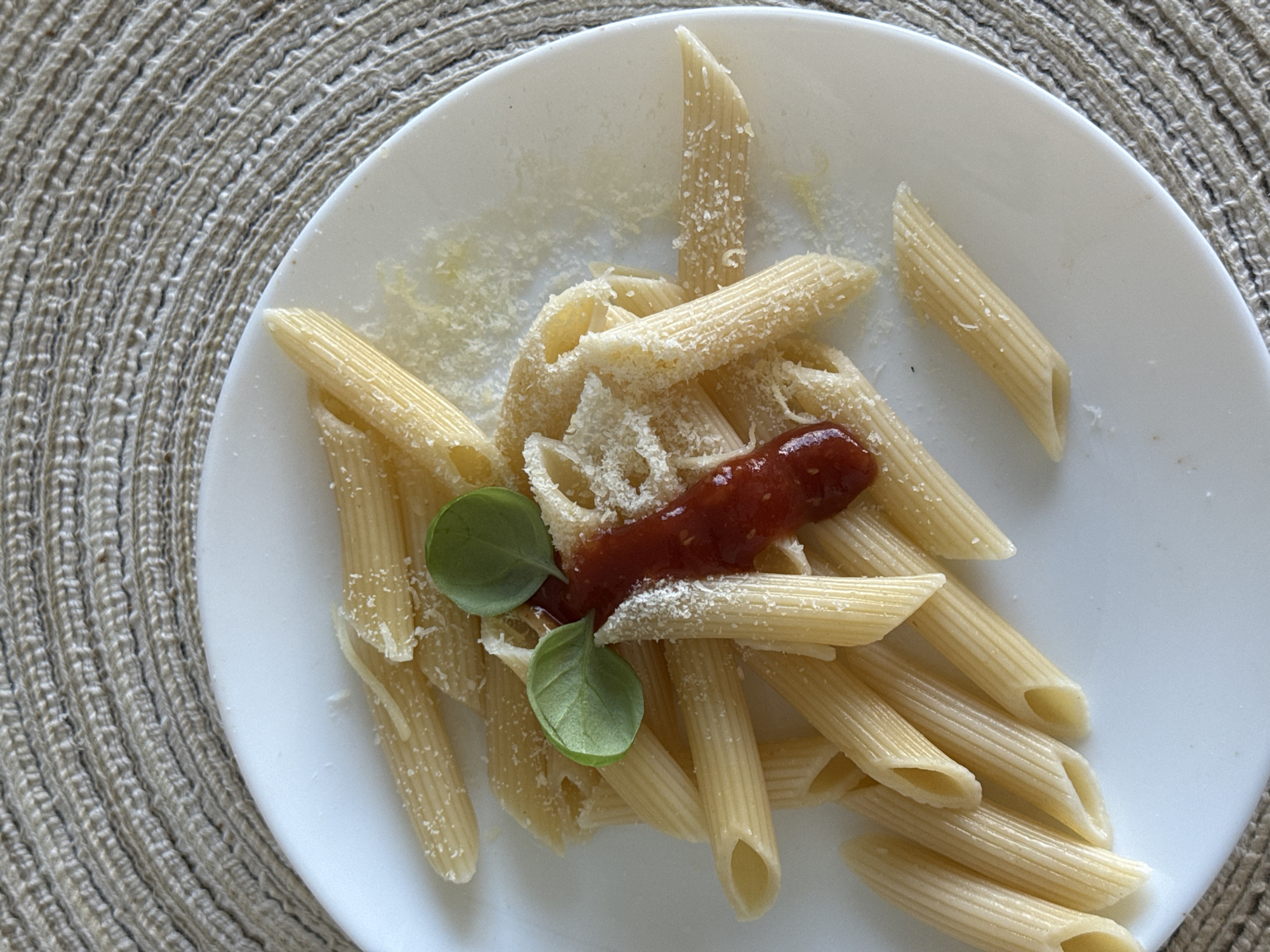 This is a close-up, overhead shot of a plate of penne pasta served with a dollop of red sauce and topped with grated cheese and basil leaves. The pasta is arranged artfully on a white plate, which is set on a circular, woven placemat. The background is slightly out of focus, but the texture of the placemat is clearly visible. The lighting is natural, creating soft shadows. There are no people visible in the image, and no indication of the specific location being in Гамово, Russia.