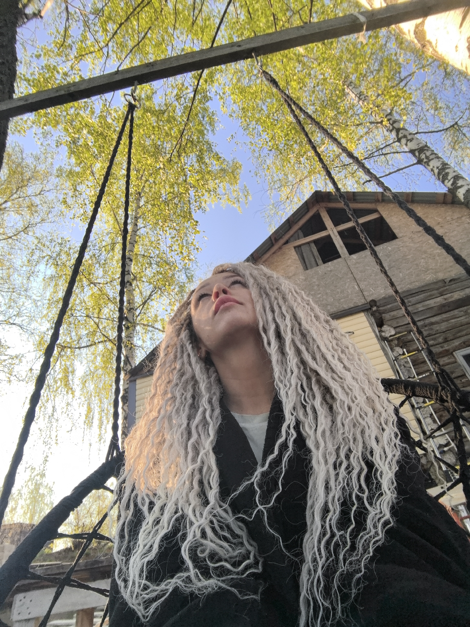 A low-angle shot captures a woman with long, light-colored, curly hair looking up. She is wearing a dark coat or jacket. The photo appears to be taken outdoors, possibly in a park or garden. The environment includes trees with green leaves and a house with light wood paneling. Rope swings frame the foreground, suggesting the woman is sitting on a swing set. The location is suggested as Noviy, Russia.