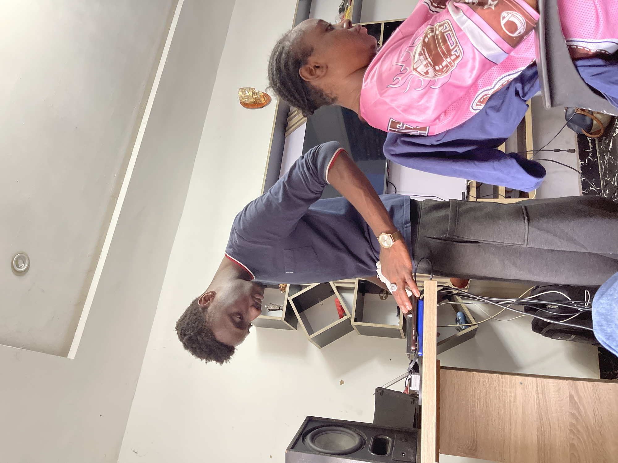 The image depicts two individuals indoors. A younger man in a dark blue shirt and grey pants is standing near an audio setup. An older woman wearing a pink and purple outfit is nearby. The setting includes a wall with geometric shelving. Cables and electronic equipment are visible. The location is potentially in Abuja, Nigeria. The specific actions or interactions between the individuals are not clear from the image.