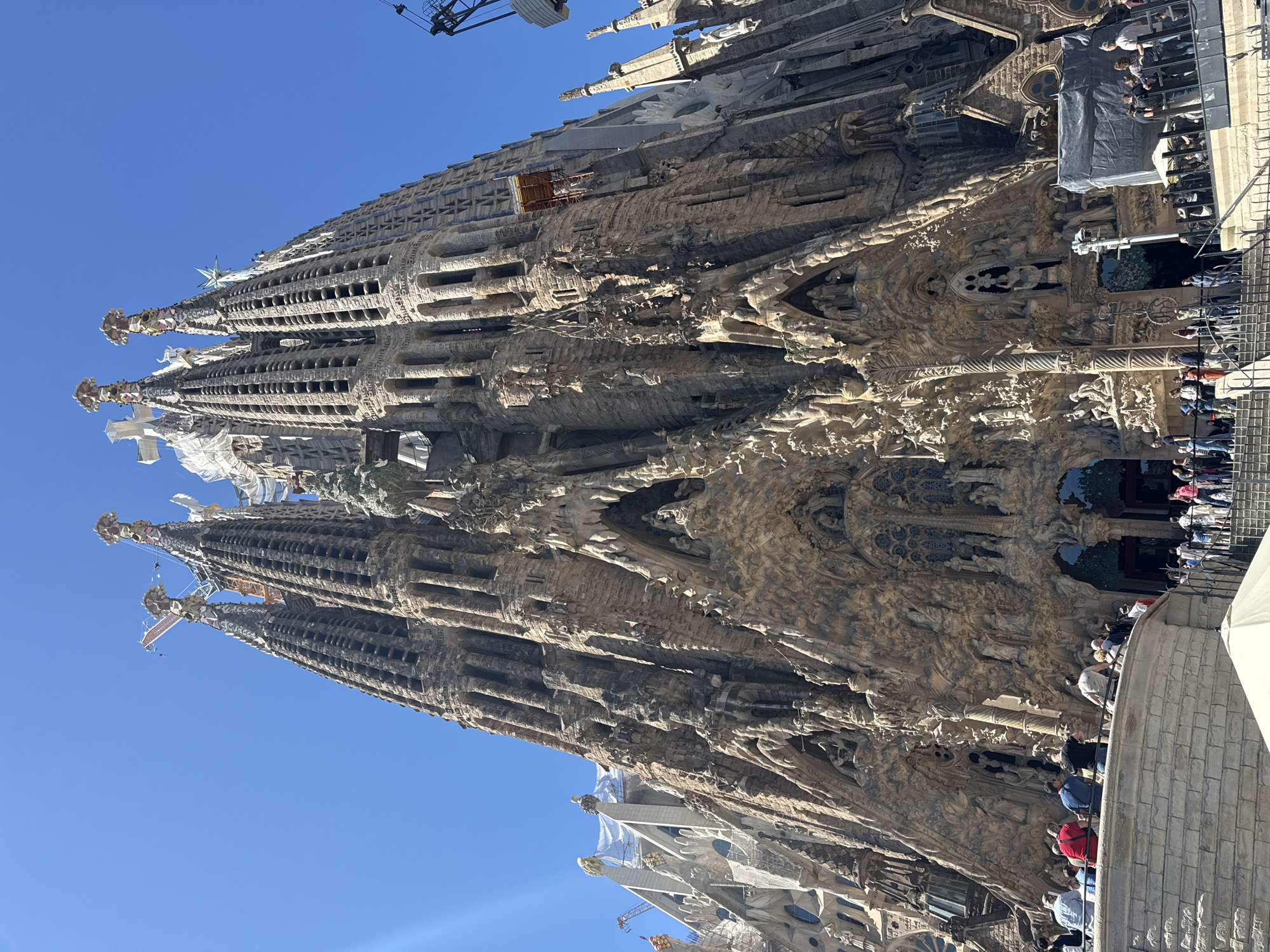 The image depicts the Sagrada Familia basilica in Barcelona, Spain. The exterior facade, showcasing intricate sculptural details and towering spires against a clear blue sky, is visible. A construction crane is present near the top of the basilica. People are positioned along a low wall near the base of the structure.