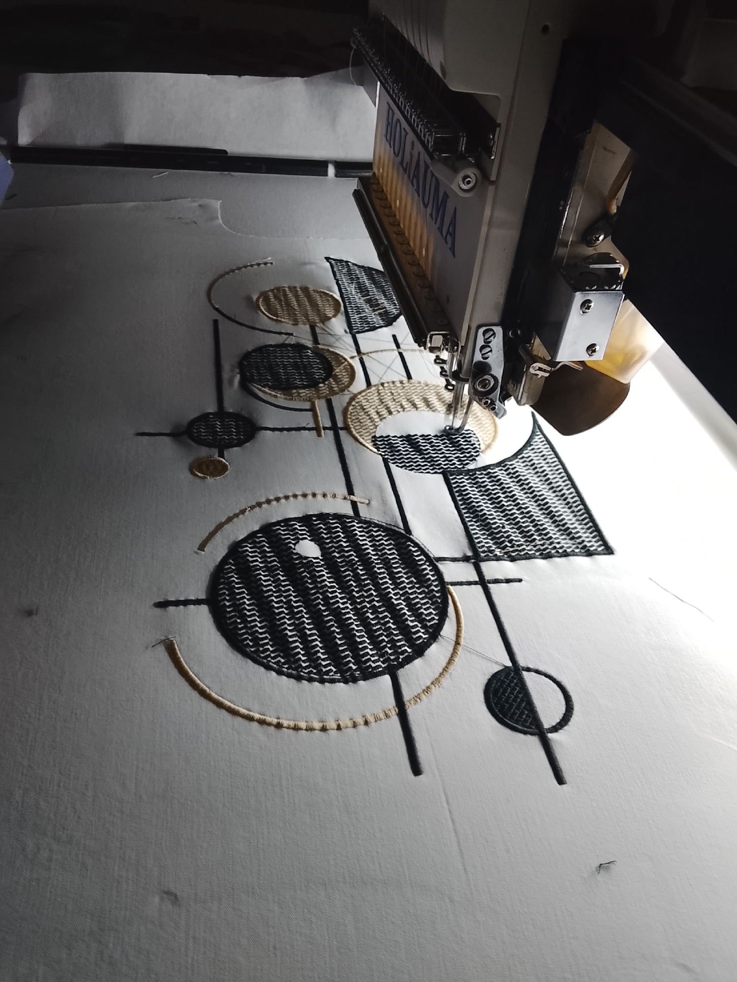 The image depicts an industrial embroidery machine in operation, stitching a geometric pattern onto a white fabric. The machine, labeled "HOLIAUMA," is positioned at an angle, with its needle mechanism visible. The fabric is stretched taut on a hoop, and the partially completed design features circles, lines, and textured fills in black and tan threads. The background is dark, suggesting an indoor environment, possibly a workshop or factory. There are no people visible in the image. The scene is devoid of any specific geographic markers, and therefore its location in Rumuepirikom, Nigeria cannot be determined.