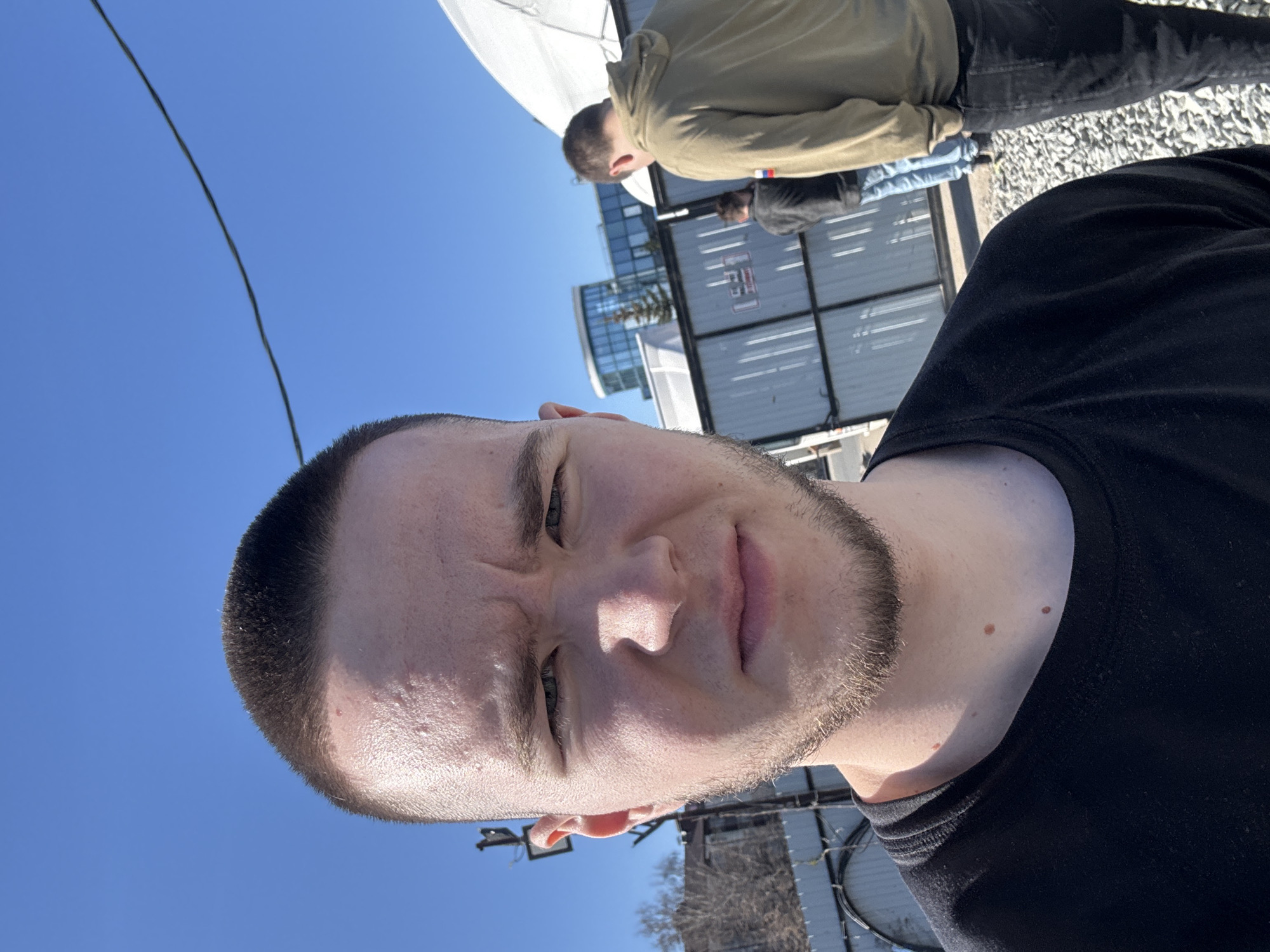 A selfie-style photograph shows a young man with closely cropped dark hair and a short beard, wearing a black shirt. He is looking directly at the camera with a slightly furrowed brow. Behind him, two other individuals are visible. One wears a tan jacket and black pants, while the other is dressed in dark clothing. The setting appears to be an outdoor area with metal fencing and a building structure featuring grid-like panels. The background includes a clear blue sky and some bare trees. There is insufficient detail in the image to positively identify the location as Kazan, Russia, or provide any other specific details about the environment.