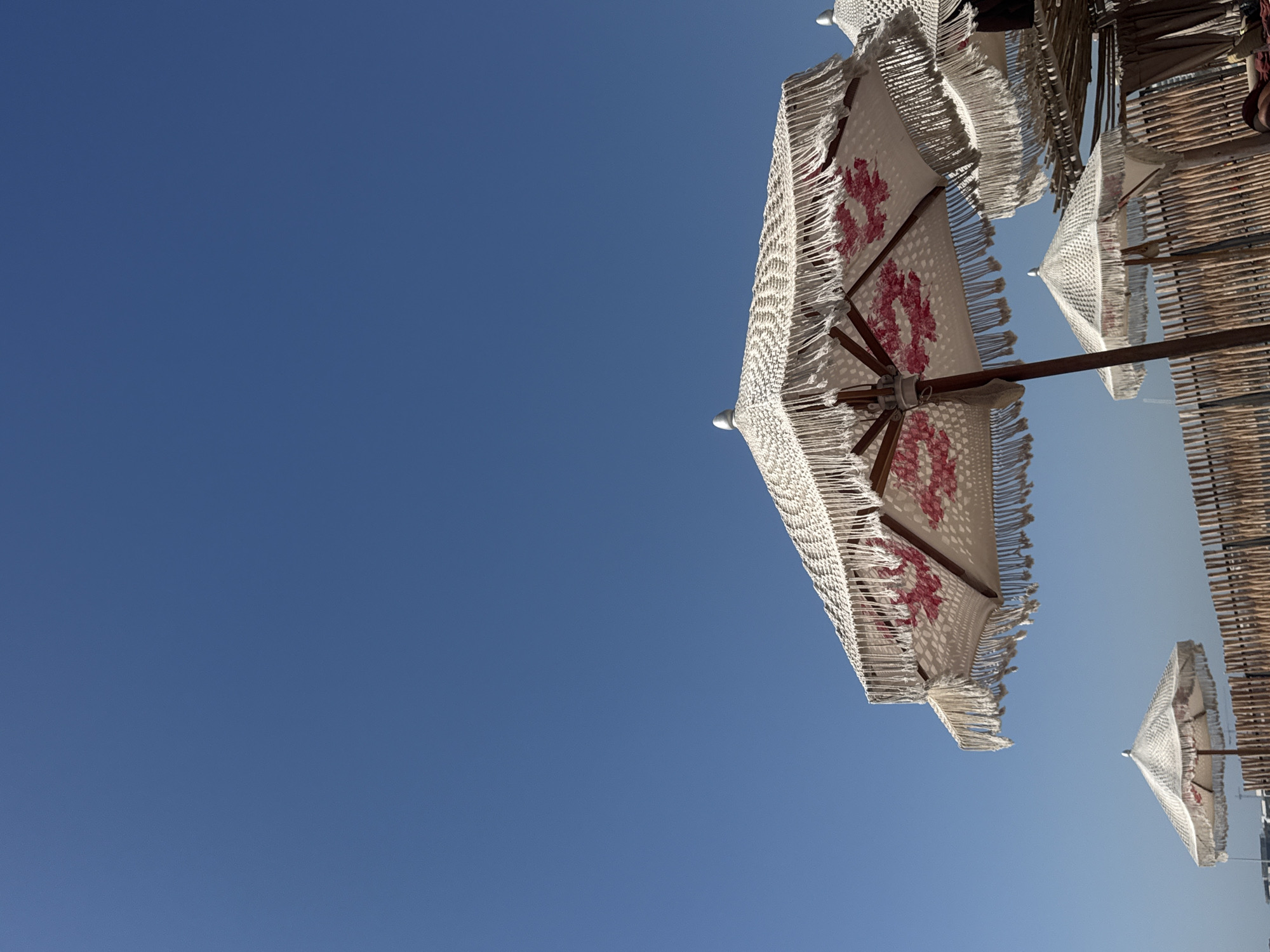 The photograph depicts a view of beach umbrellas against a clear, bright blue sky. The umbrellas are white with decorative fringes and pink floral patterns. The scene is located in Dubai, United Arab Emirates. The photo is taken from a low angle, looking up at the umbrellas. Some of the umbrellas appear to be attached to a bamboo structure. There are no people visible in the image.
