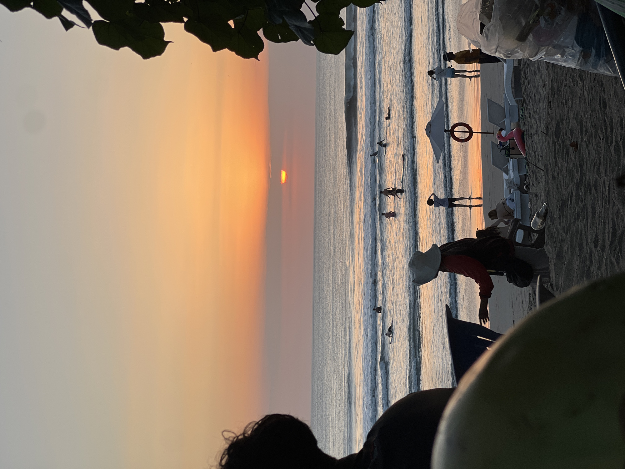 An outdoor scene at sunset captures the Kuta beach in Indonesia, with the sun partially obscured by a band of clouds as it sets over the ocean. The sky transitions from a pale blue at the top to hues of orange and pink closer to the horizon. Gentle waves roll onto the sandy shore, where several surfers are visible in the water, some paddling and others riding waves.
On the beach, a person wearing a wide-brimmed hat stands in the foreground, facing the ocean. Behind them, beach chairs and an umbrella are set up. A bright pink inflatable flamingo lies on the sand near the chairs. To the right, a large, clear plastic bag filled with trash sits on the beach, with a few bottles scattered nearby. The scene is framed by dark foliage on the right side and the silhouette of a person in the lower left corner.