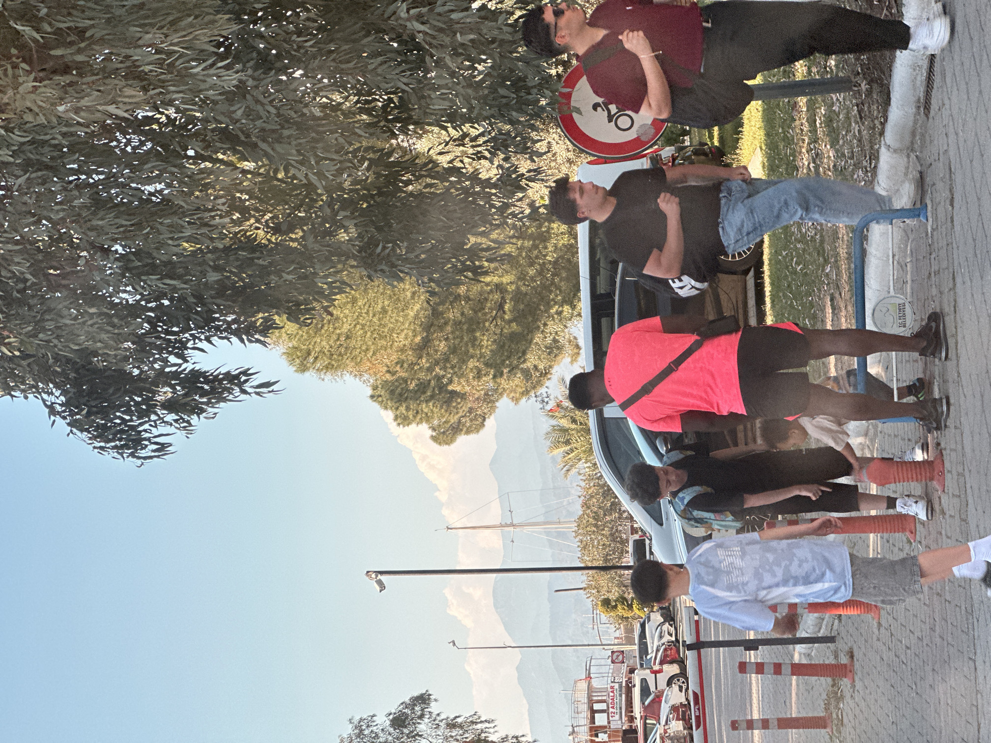 The image, taken in Fethiye, Turkey, depicts a vibrant outdoor street scene under a clear blue sky. In the foreground, several individuals are visible. On the left, two men stand beneath the canopy of a large, leafy tree. One wears a maroon shirt and dark trousers, while the other is in a black t-shirt and blue jeans, leaning against a low blue metal barrier. Behind them is a circular "no bicycles" sign.
Towards the mid-ground, a person with dark skin wears a bright neon pink/orange top and dark shorts, standing next to a white van. Further right and in the foreground, several other people, including children and young adults, are gathered near a series of orange traffic cones. One young person wears a light blue patterned t-shirt and grey shorts.
The background features distant mountains under a clear sky, with patches of green foliage and a visible sailboat mast, hinting at a coastal location. Sunlight suggests a bright day, likely morning or late afternoon, casting some shadows.
Visible text includes:
* "TC FETHİYE BELEDİYESİ" on a white sign, indicating "Republic of Turkey Fethiye Municipality."
* "12 ADALAR VE ÖLÜDENİZ" on another sign, referencing "12 Islands and Oludeniz," popular local tourist destinations.
* The "no bicycles" symbol on a road sign.
The scene conveys a casual public gathering or waiting area, possibly near a transportation hub or a tourist attraction in Fethiye.