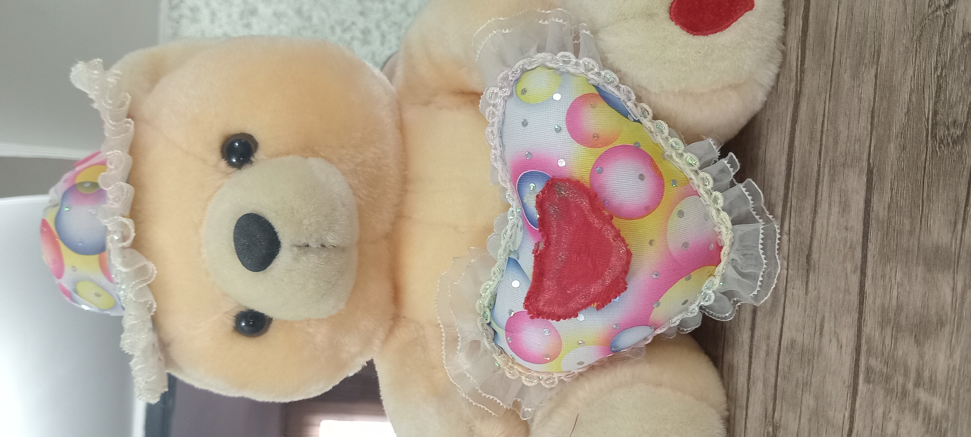 A close-up, vertical, eye-level shot shows the upper body of a light beige teddy bear. The bear is wearing a colorful, polka-dot hat with a white, ruffled brim. Its front is adorned with a heart-shaped pillow that features a pattern of colorful circles and sequins. A small, red fabric heart is attached to the center of the pillow. The bear's paws are visible, resting on a surface that appears to be a wooden table with a distressed, grey-brown finish. The background is out of focus, showing a mottled, light-colored wall and a hint of a darker area to the left.