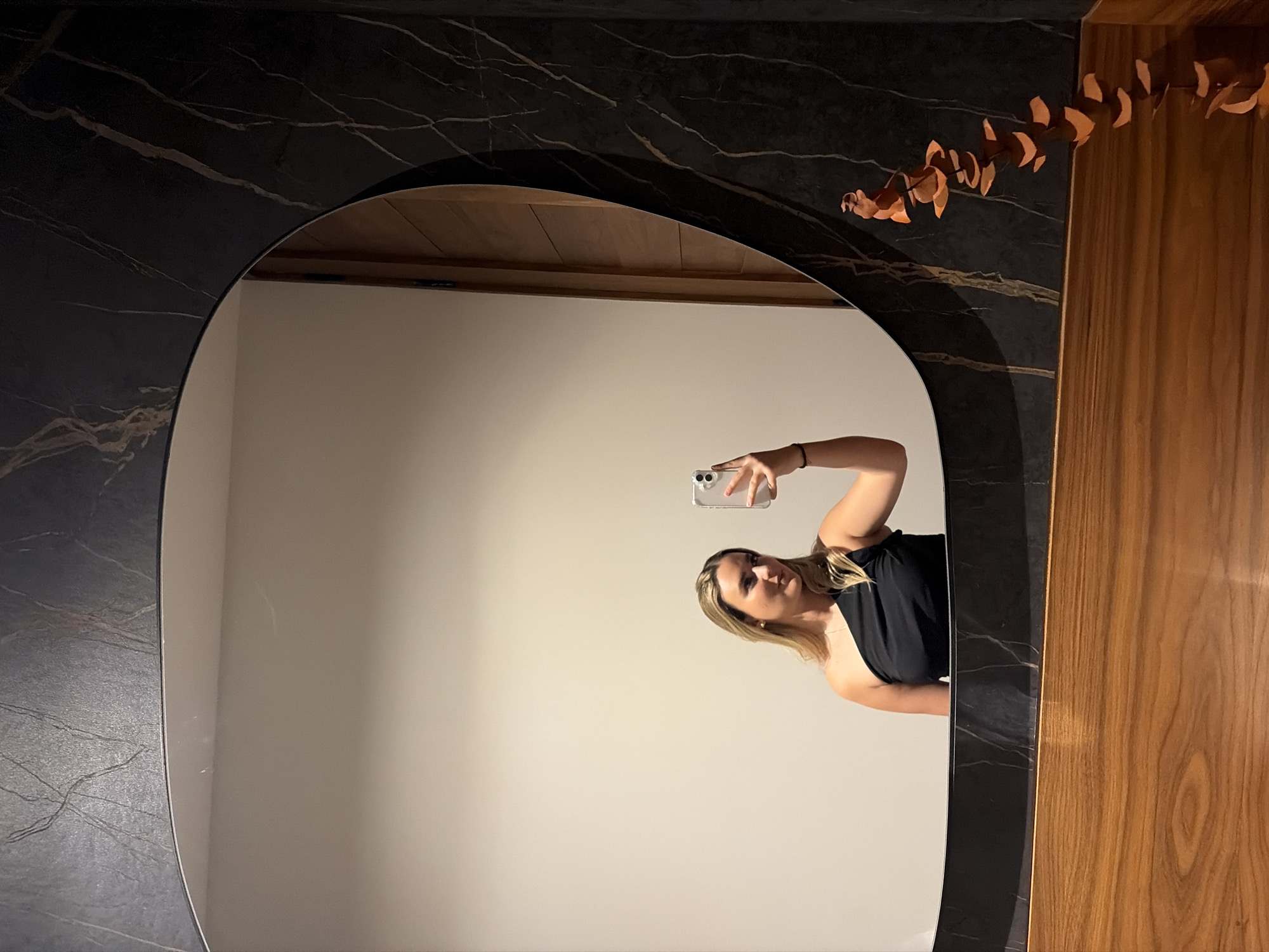 A woman is taking a selfie in a mirror. The mirror has a dark gray, marble-like frame. To the right of the mirror is a wooden panel with an orange eucalyptus branch. The background is a plain, off-white wall. The woman is fair-skinned with blonde hair and is wearing a black top. She is holding a smartphone in her right hand, presumably to take the picture. The reflection shows a portion of a wooden ceiling. The location is not discernible.