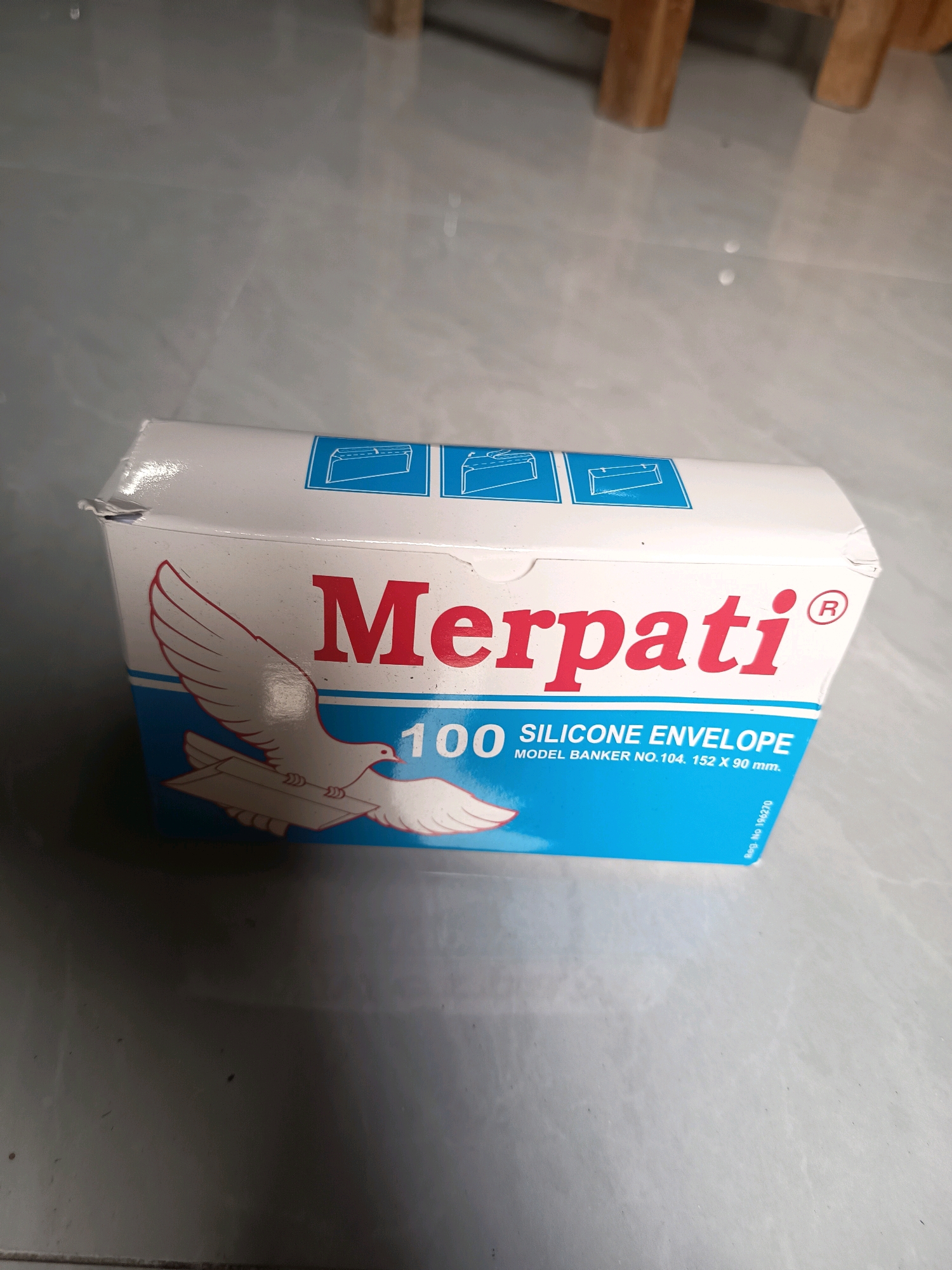 A top-down, close-up, eye-level shot shows a white and blue box of "Merpati" brand silicone envelopes. The box has a red logo of the word "Merpati" and a drawing of a white dove carrying an envelope. Text on the box indicates "100 SILICONE ENVELOPE MODEL BANKER NO. 104. 152 X 90 mm." and "Reg. No. 196270". The box is positioned on a gray, tiled surface. The background is out of focus and appears to be a light gray floor with some wooden furniture legs visible at the top. The scene is indoors, suggesting it is a retail setting or a home.