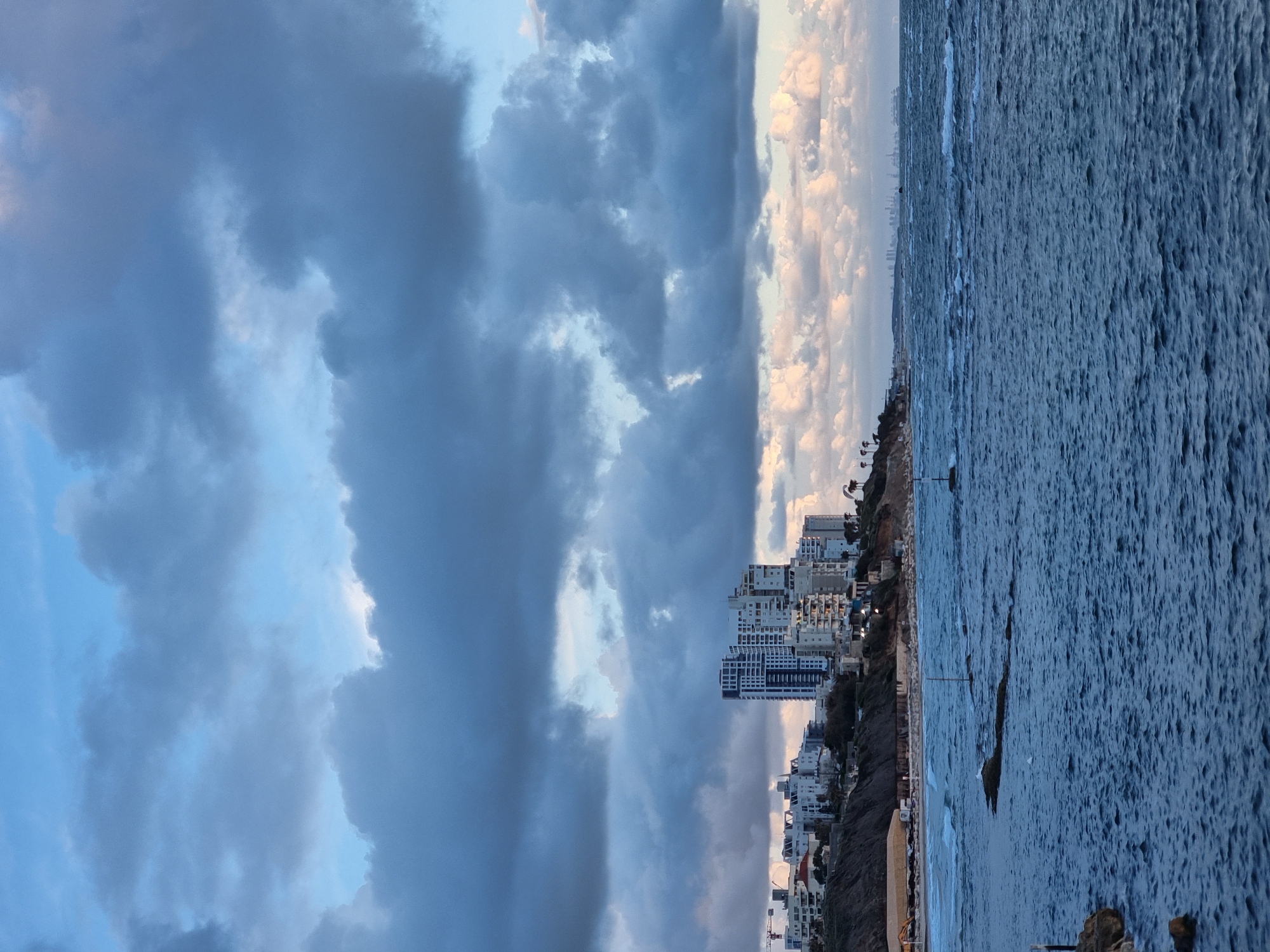 The image displays a coastal panorama in Tel-Aviv, Israel, under a partially overcast sky. The upper two-thirds of the frame are filled with dense, dark grey cumulus clouds, interspersed with patches of light blue sky and a brighter, light-toned glow on the right horizon.
The lower third of the image is dominated by the Mediterranean Sea, exhibiting dark blue-grey, choppy water with visible waves and whitecaps. Several dark, elongated rock formations are partially submerged in the mid-distance. A thin, vertical pole is present in the water towards the center-right, and another shorter pole is visible near submerged rocks in the lower-left foreground.
The coastline extends from the left to the mid-right, featuring a steep, vegetated cliff. A narrow strip of sandy beach lies at the base of the cliff, where several light-colored, temporary structures, possibly tents, are present. A yellow excavator or similar construction equipment is visible on the far left portion of the beach. Faint human figures are discernible on the beach.
Perched atop and along the cliff are numerous multi-story residential buildings, primarily white or light-colored. One prominent building in the center of the cliff area features a distinctive dark blue-grey vertical section on its facade. A large construction crane stands on the far left, above the cliff, indicating ongoing development. Palm trees are scattered along the cliff's edge. In the far distance on the right horizon, a faint silhouette of a larger urban skyline or port structures is visible.