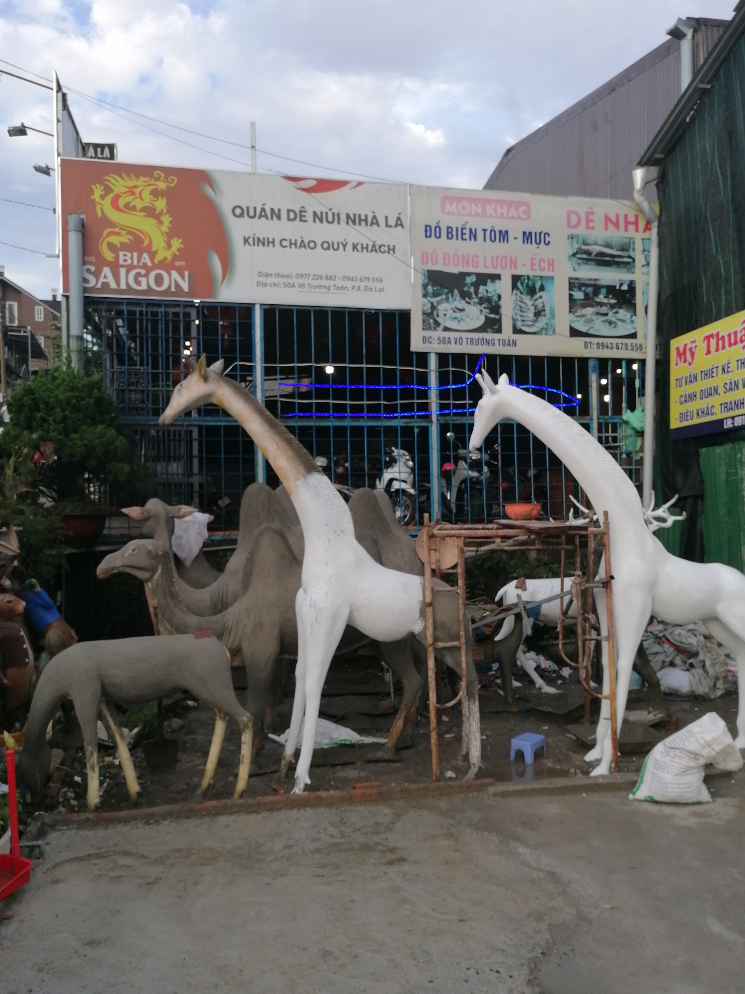 The image shows a storefront with a large sign for "QUÁN DẺ NÚI NHÀ LÁ" which translates to "Leaf House Mountain Restaurant" in Lâm Viên - Đà Lạt, Vietnam. The restaurant appears to specialize in seafood, with "MÓN KHÁC" (other dishes) including "ĐỒ BIỂN TÔM - MỰC" (shrimp and squid seafood) and "ĐỒ ĐỒNG LƯƠN - ÉCH" (eel and frog dishes). The address is listed as "50A Võ Trường Toản, P.8, Đà Lạt".
In front of the storefront, there are several large, unpainted animal sculptures, including a giraffe, a camel, and a deer. The sculptures are in various stages of completion, with some appearing to be made of fiberglass or concrete. The environment is a street scene with a cloudy sky. There are no people clearly visible in the image, though there are some motorcycles behind a metal fence. The overall impression is of a business that sells or displays decorative animal figures.