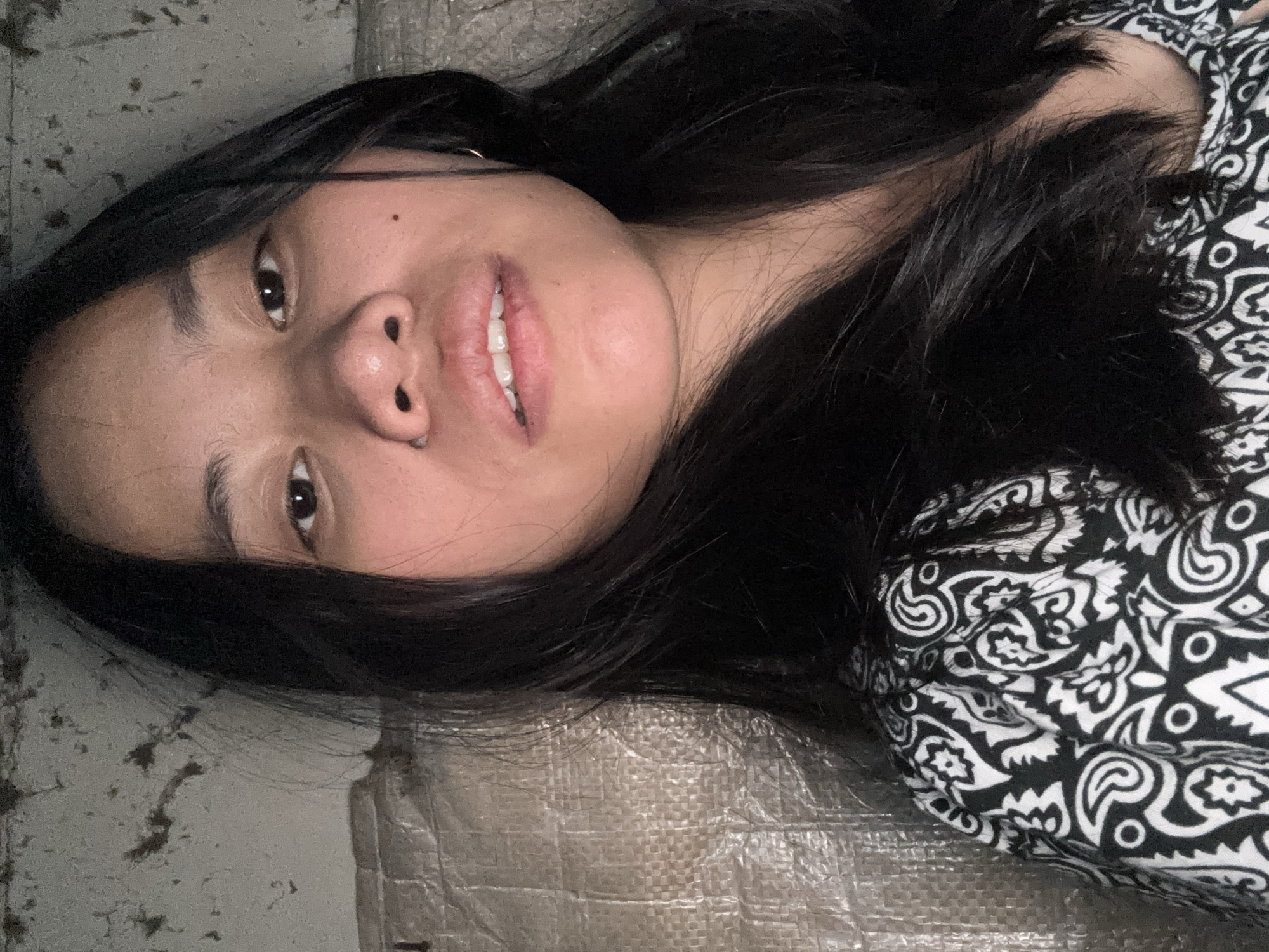 A young woman with long, dark hair is shown in a close-up portrait. She is wearing a black and white patterned top. Her gaze is directed towards the viewer, and her lips are slightly parted, revealing her teeth. She has a mole on her right cheek. The background appears to be a textured wall and a woven material. The image was taken in East Jakarta, Indonesia.