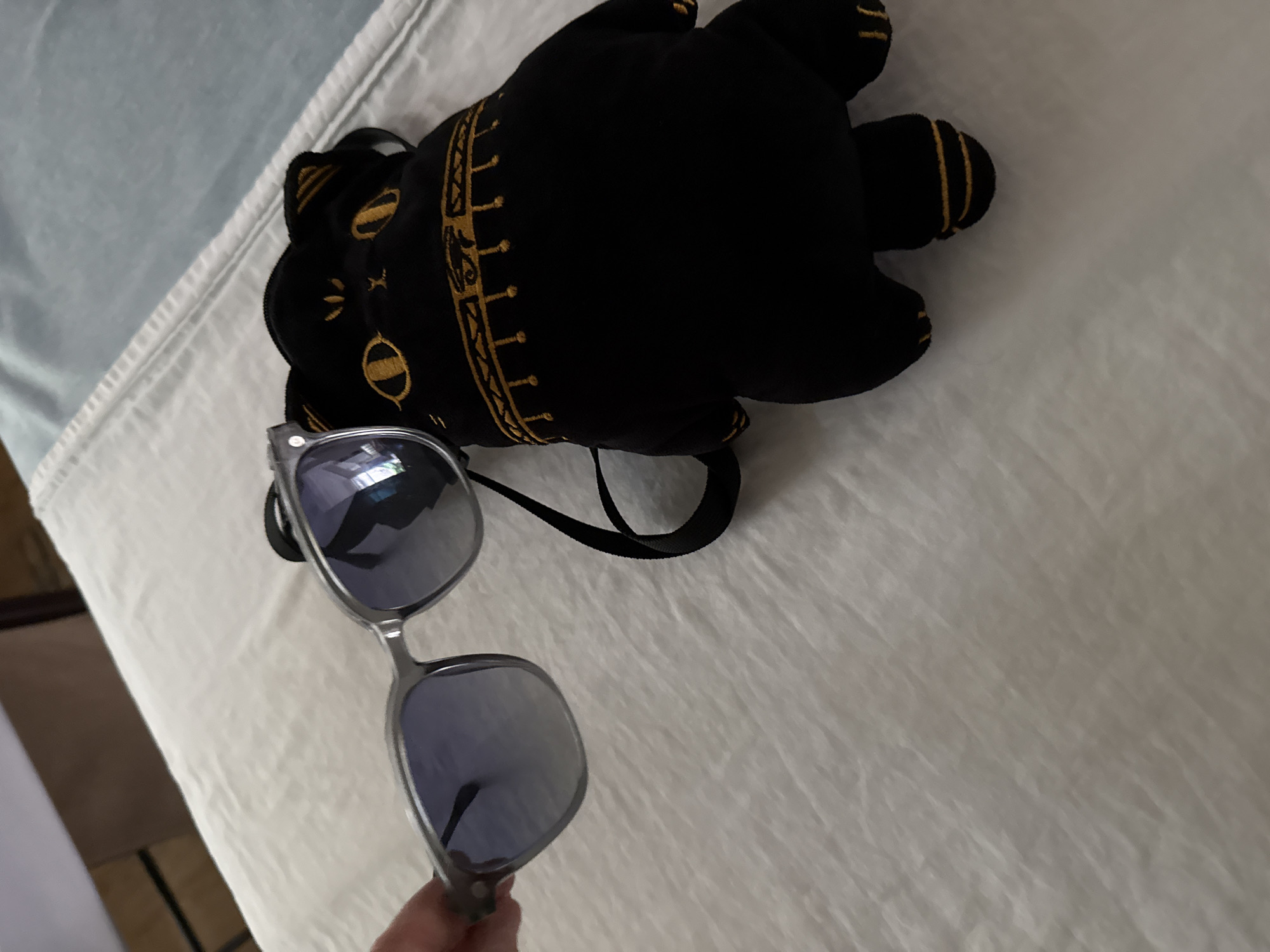In Huizhou, China, on a white, textured surface that appears to be a bedspread, a person's hand is holding a pair of translucent gray-framed sunglasses with blue lenses. To the right of the sunglasses lies a black plush toy shaped like a cat's head. The cat toy has embroidered gold eyes and decorative gold trim around its face and what appear to be paws. A black strap is attached to the cat toy, suggesting it might be a bag or backpack. The background is out of focus, showing a blurred light blue surface and a brown object.
