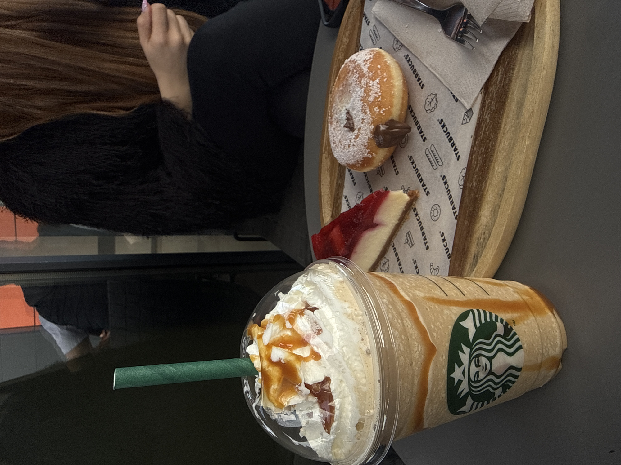 This image depicts a spread of Starbucks food and drinks on a grey table. A large caramel Frappuccino with whipped cream and a green straw is positioned in the foreground. Behind it, a wooden tray holds a powdered donut with a chocolate filling, a slice of cheesecake with strawberry topping, a napkin, and a fork. Starbucks branded paper lines the tray. To the side, a person with long brown hair and wearing a black fuzzy sweater is seated. The setting appears to be inside a Starbucks, with a window showing a reflection of people and buildings. Although the location is a Starbucks, the specific city of Plovdiv, Bulgaria, cannot be determined from the image.