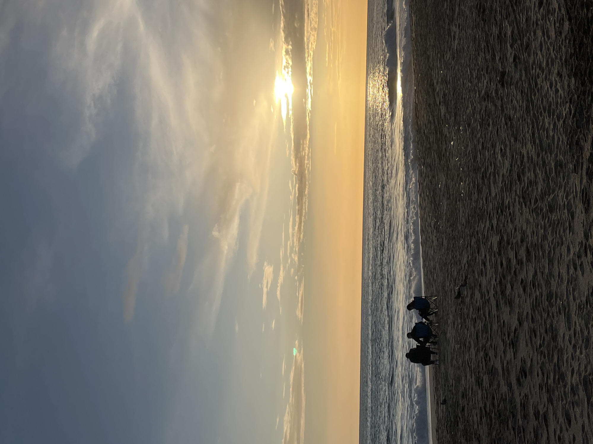 Here is a forensic description of the image:
The image depicts a beach scene, likely taken at sunset or sunrise given the low angle of the sun and the color of the sky. The location is most likely Alanya, Turkey.
The left half of the image shows the sky, which transitions from a light blue at the bottom to a yellow and orange hue near the horizon. There are scattered clouds. The sun is low on the horizon, and its light is reflected on the ocean.
The right half of the image shows the beach and the ocean. The sand is dark in color. Waves are visible, breaking on the shore.
There are three figures sitting on beach chairs near the shoreline. The people and chairs are silhouetted against the bright light of the sunset/sunrise. The figures appear to be facing the ocean. There are no visible interactions between the figures.