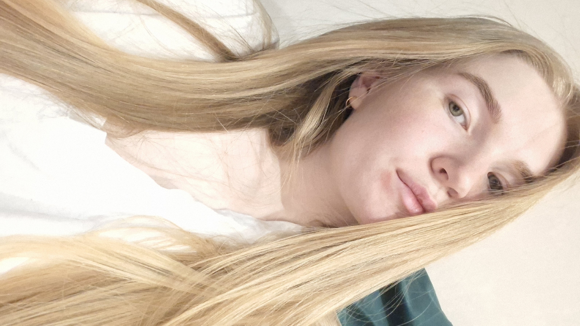 A close-up, softly-lit selfie of a fair-skinned young woman with long, straight blonde hair. The hair is partially covering her face and cascades across a white surface, possibly a bed or pillow. She has light eyebrows, green eyes, and slightly parted lips. A small gold hoop earring is visible in her left ear. Part of a dark green garment can be seen in the lower right corner. The location in the city of Chaikovsky, Russia, cannot be determined from the image.