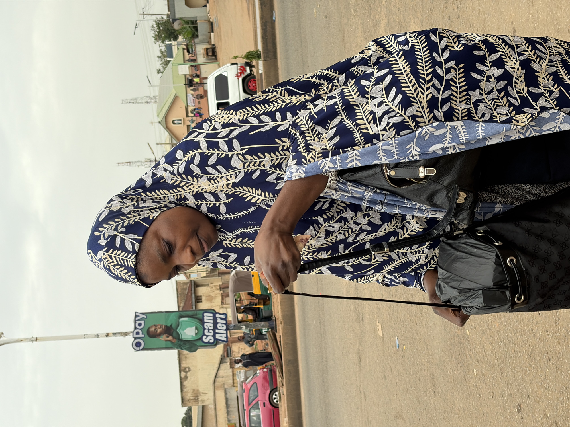 A woman is standing on a paved surface, looking down at her hands which are holding a small object and a black bag. She is wearing a dark blue hijab with a white and gold leaf pattern and a matching patterned outer garment. She has a black handbag slung over her shoulder and is holding a black umbrella in another bag in her right hand. The background shows a street with several vehicles, including a red car, a white van, and a three-wheeled auto-rickshaw. Buildings and utility poles are visible behind the street, and there is a "OPay Scam Alert" sign on a pole. The sky is overcast.