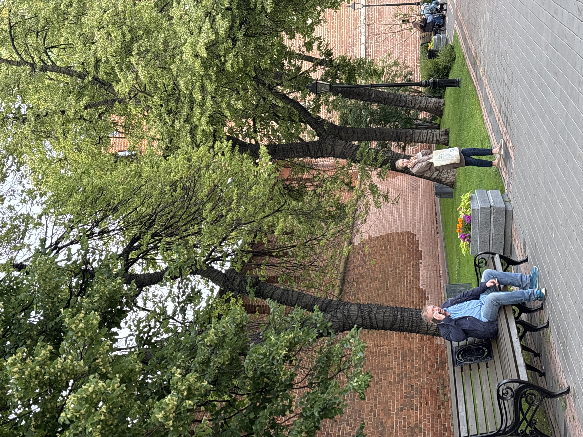 In Nizhny Novgorod, Russia, on a cloudy day, a man is seated on a park bench, speaking on his mobile phone. He is wearing a blue collared shirt, a dark blue jacket, and blue jeans. To his right, a woman stands holding a white shopping bag and also appears to be on her phone. The park features a brick wall in the background, with mature trees providing shade over a grassy area and a paved walkway. A streetlamp is visible behind the woman. In the distance, another bench is occupied by two individuals. The scene takes place in an outdoor urban park.