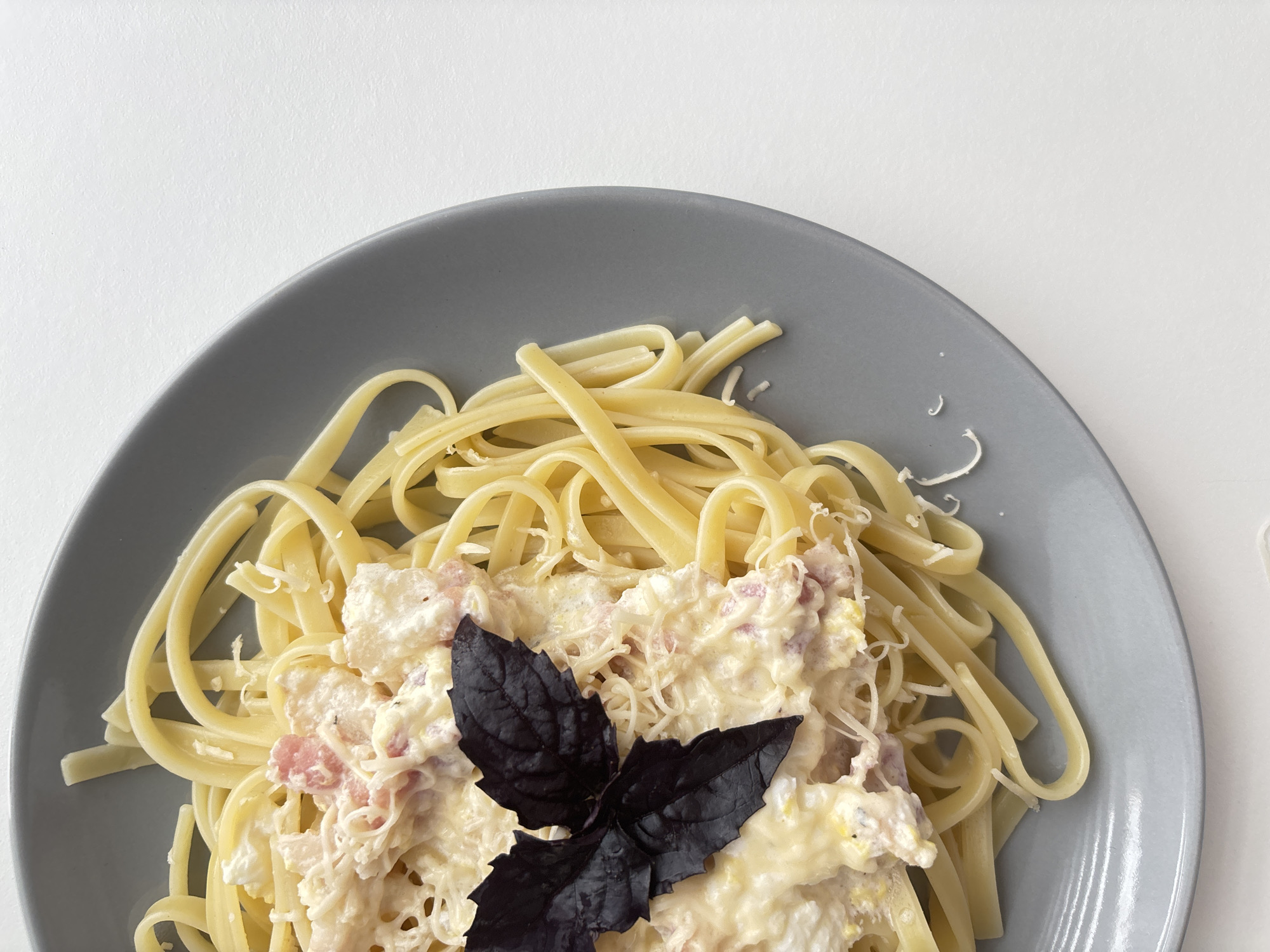 A close-up, overhead shot features a portion of a grey plate filled with fettuccine pasta topped with a creamy sauce containing pieces of bacon and shredded cheese. A dark purple basil leaf is placed on top of the pasta. The plate rests on a white surface, with the background being a solid white. There are no people or discernible city landmarks visible.