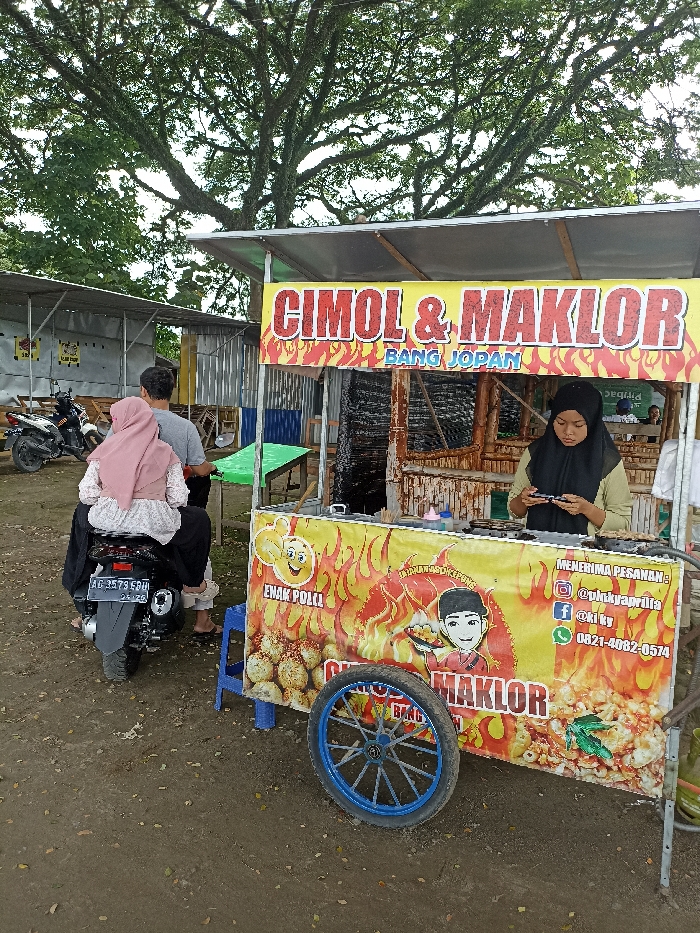 This outdoor daytime photograph captures a vibrant food stall in Kepung, Indonesia, against a backdrop of large trees and simple structures. The central subject is a food cart, operated by a woman in a black hijab and light-colored top, who is seen looking down, possibly engaged in food preparation or checking her phone. The cart's main banner is yellow with red flame accents, boldly displaying "CIMOL & MAKLOR" and "BANG JOPAN." The lower banner features cartoon imagery, illustrations of the snacks, and text that reads "ENAK POLKL" and "MENERIMA PESANAN:" (Accepting Orders), along with social media handles "@phakyaprilia," "f@kiky," and a phone number "0821-4082-0574."
To the left, a black scooter is parked on the dirt ground. A person wearing a pink hijab and a white patterned top sits on the scooter's rear, while another individual stands nearby, partially obscured. The scooter's license plate, "AG 3579 EDU," is visible. The setting is rustic, with a dirt ground and corrugated metal shacks in the background. Overhead, a large tree with abundant, gnarled branches and green foliage provides a natural canopy, suggesting a somewhat rural or natural environment for this local food business. The lighting is soft and diffused, indicating an overcast day or indirect sunlight.