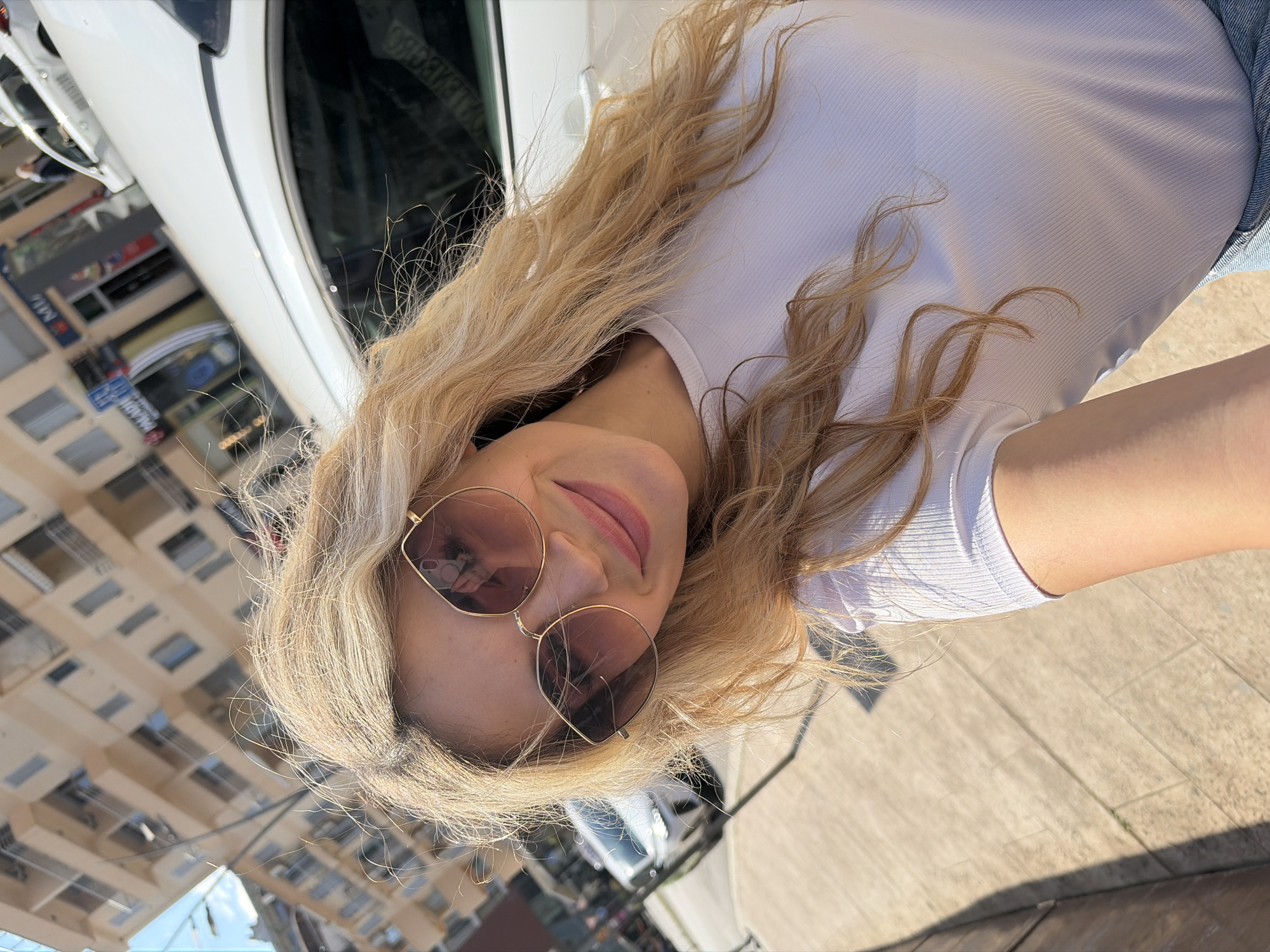 A woman with long, blonde wavy hair poses for a selfie outdoors on a sunny day. She wears sunglasses with round frames and a white, short-sleeved shirt. She is near a white vehicle with reflective windows. In the background, a cityscape is visible with buildings under a blue sky. Buildings behind her suggest a location within the urban environment of Budva, Montenegro. The ground appears to be concrete or pavement.