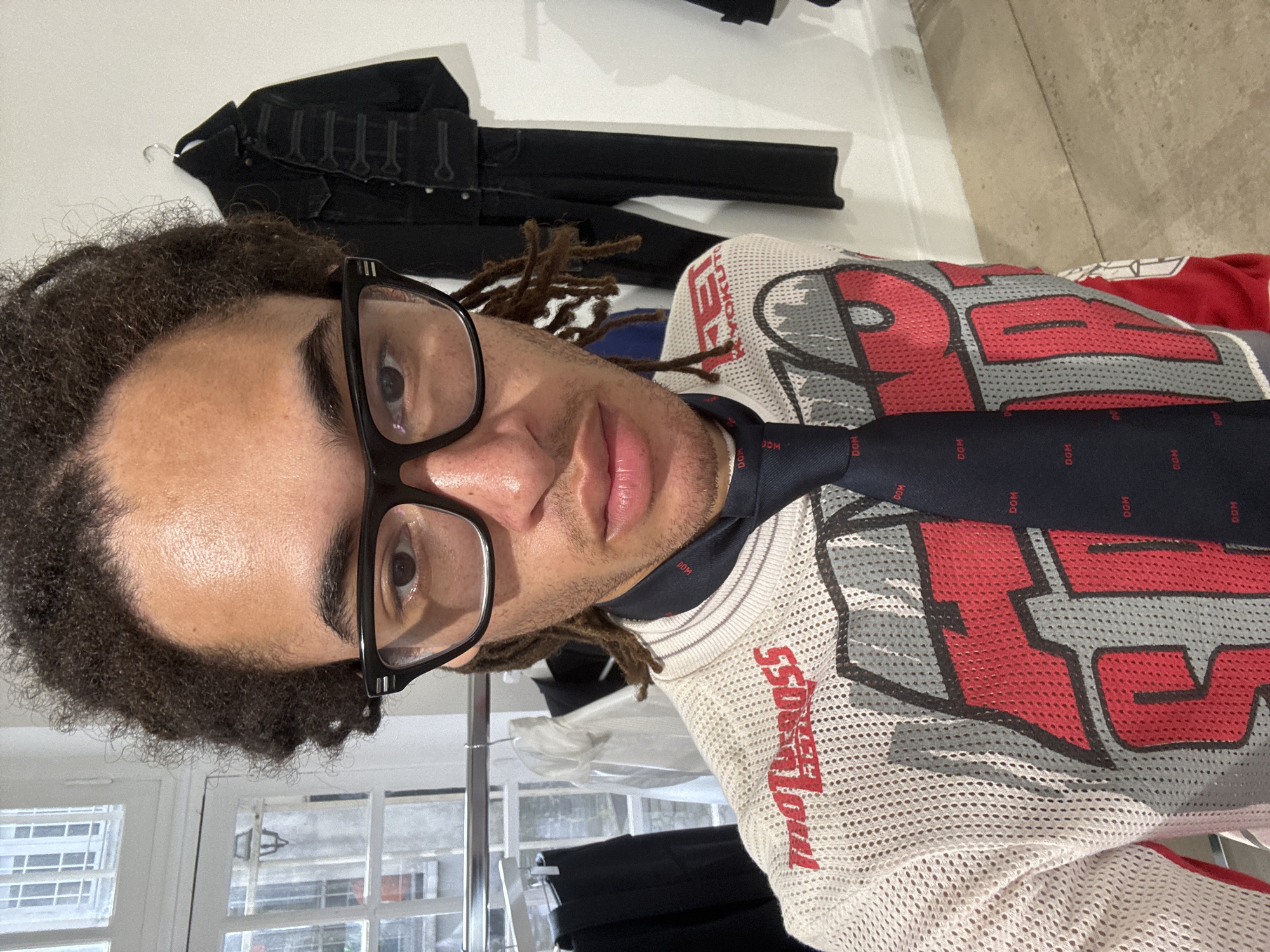 A person with dark curly hair and dreadlocks, wearing black-framed glasses and a white mesh shirt with red and gray lettering and graphics, stands indoors in an area that appears to be a clothing store or studio. They are wearing a dark tie with repeated "DOM" text. Behind the person, on a white wall, hangs a dark garment with button or strap details. In the lower left, a white-framed window reveals a view of buildings outside, suggesting an urban setting. Based on the architecture and the style of the shop, the location may be Paris, France. Clothing racks and various garments are visible in the background.