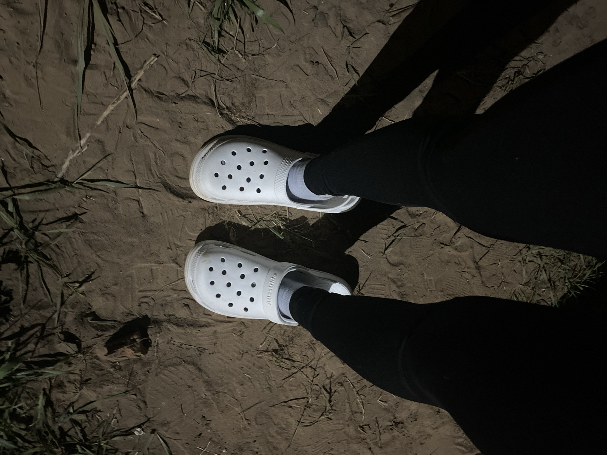 A person wearing black leggings and white Croc-style shoes with white socks stands on a dirt path with some grass and small twigs. The scene appears to be illuminated by a single light source, casting shadows on the legs and the ground. The location is unspecified.