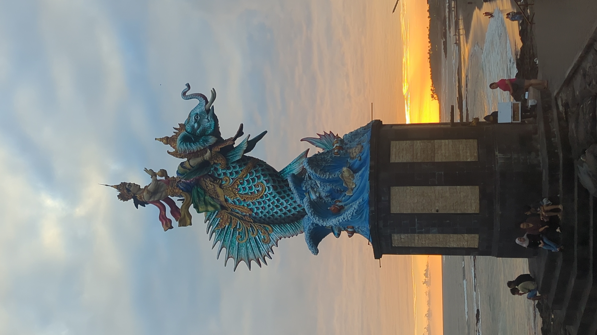 A statue of a Hindu deity riding a mythical creature stands on a pedestal overlooking the ocean at sunset. The deity, adorned with a crown, is depicted with a serene expression, holding an object in one hand and making a gesture with the other. The creature is a fusion of a fish and an elephant, with colorful scales and an elephant's head. The base of the statue is decorated with crashing waves and various sea creatures. Several people are seated on steps in the foreground, observing the view. The background features a beach with a few people walking, and the sun is setting over the horizon, casting a warm glow on the water and sky. The statue appears to be located in a public area, possibly a tourist attraction in Mengwi, Indonesia.