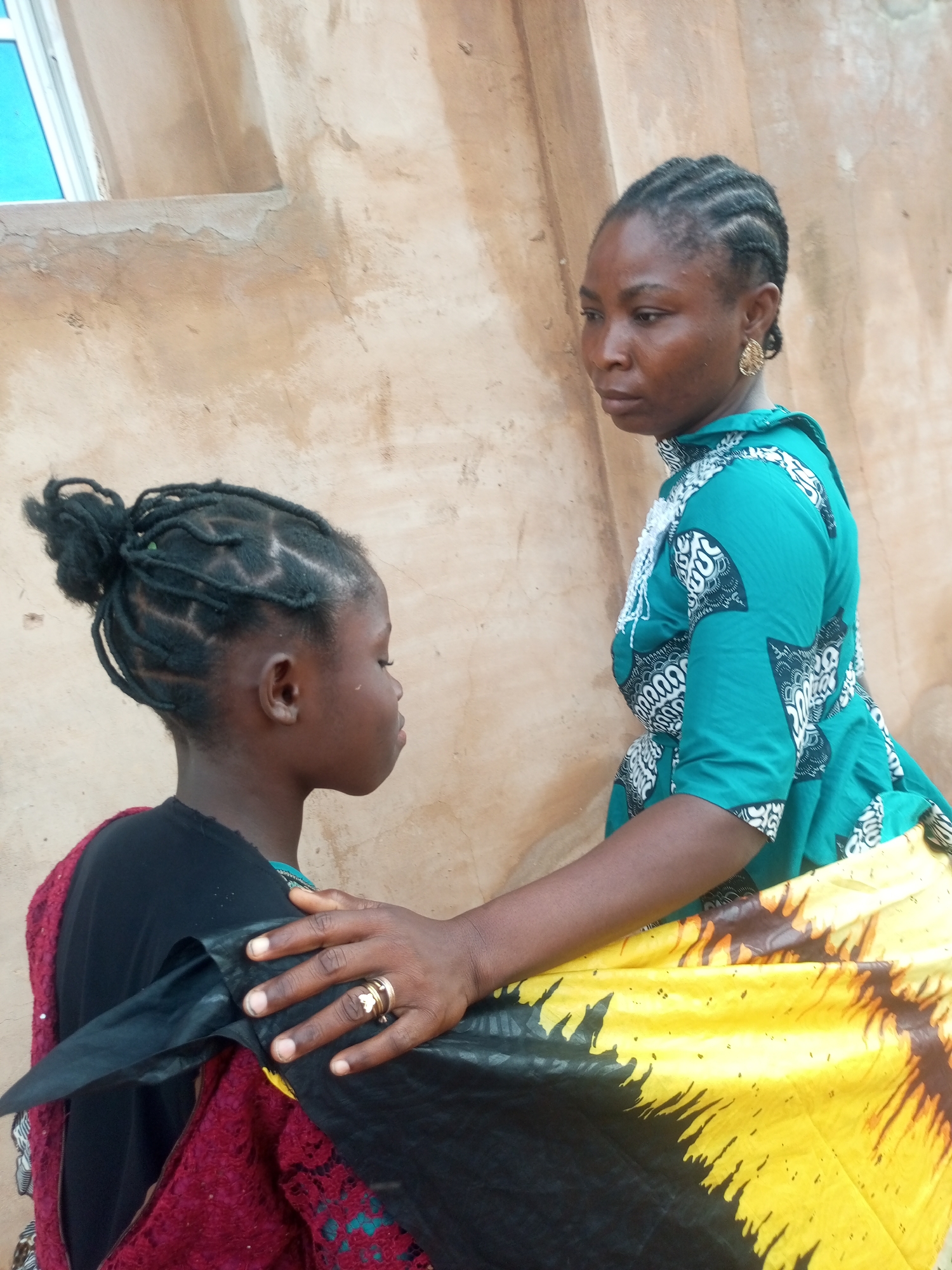 In an outdoor environment, two Black females are positioned in front of a textured, tan-colored wall, likely in Tempure, Nigeria. The female on the right, who appears older, is wearing a teal green top with a black and white patterned print. She has her hair braided in a cornrow style and is wearing a large, gold, teardrop-shaped earring. Her right hand is gently placed on the shoulder of the younger female.
The younger female, on the left, is facing away from the viewer and looking to her right. She has her hair styled in intricate braids that are gathered into a bun at the back of her head. She is wearing a black top layered over a burgundy, lace-trimmed garment. A portion of a yellow and black patterned fabric is draped across her and the older female's laps. The older female's hand, adorned with a gold ring featuring a heart-shaped embellishment, rests on the younger female's shoulder. To the left of the frame, a small section of a light blue window or frame is visible against the wall. The lighting suggests it is daytime.