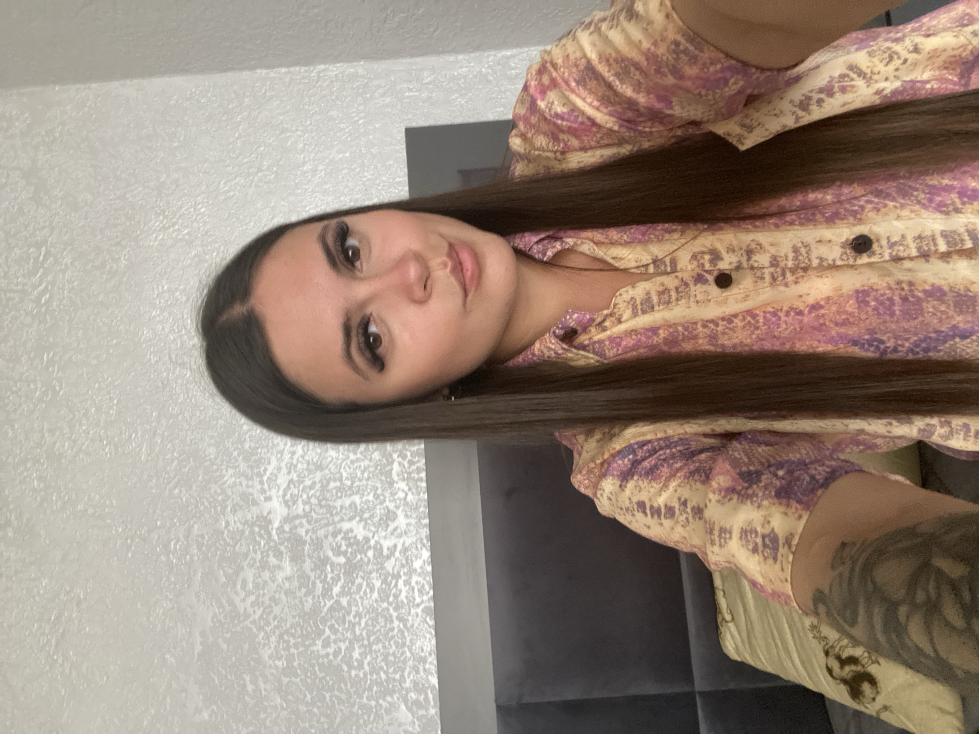 A woman with long, dark hair, wearing a collared, short-sleeved shirt with a patterned design in shades of pink and yellow, is captured in a close-up, upper-body selfie. Her head is slightly tilted, and she has on false eyelashes. She is positioned in front of a textured, light-colored wall and a dark-colored headboard, suggesting she is indoors, likely in a bedroom. A pillow with a tan and black pattern is visible to her left. There is a tattoo visible on her left arm. The scene does not provide any specific information to determine a location in the city of Chalda, Russia.
