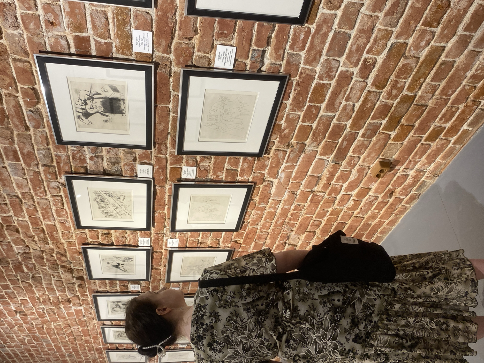 The image depicts a person, likely a woman, gazing at framed artwork displayed on a red brick wall. The artwork appears to be drawings or prints, each framed in black with a white mat. Small informational cards are placed above each frame. The person is wearing a dress with a floral pattern and a black bag is slung over their shoulder. The scene is likely in an art gallery or museum with a vaulted ceiling of exposed red brick. The informational cards are written in Russian, and they identify the artist as Marc Chagall. Based on the text on the informational cards, this scene is likely located in a gallery or museum in Russia, as it references the Russian artist Marc Chagall, in the city of Yekaterinburg.
