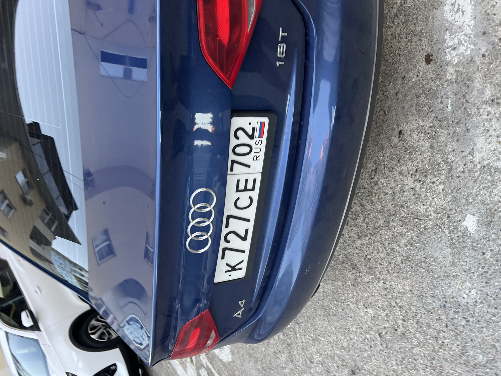 A dark blue Audi A4 sedan is shown parked outdoors, viewed from the rear at a slight angle. The car's metallic paint reflects the surrounding environment, which includes a partial view of a white car parked alongside and multi-story buildings with windows reflected on the rear window and body panels. The ground beneath the car is gray asphalt or concrete.
Notable details on the car's rear include the silver Audi rings emblem centrally located on the trunk lid. To the lower left of the trunk is the "A4" model badge, and to the upper right is the "1.8T" engine identifier. The car's red taillights are visible on both sides of the trunk.
A white Russian license plate is mounted centrally, displaying the characters "K727CE 702 RUS". The "RUS" section of the plate incorporates a small, rectangular depiction of the Russian flag (white, blue, red horizontal stripes). There is a rectangular mark above the license plate, possibly from a previously mounted plate or holder.
The image is captured during the daytime under clear weather conditions. No people are visible in the scene. The presence of the Russian license plate confirms the geographical context as Russia, with the provided information specifying Sochi.