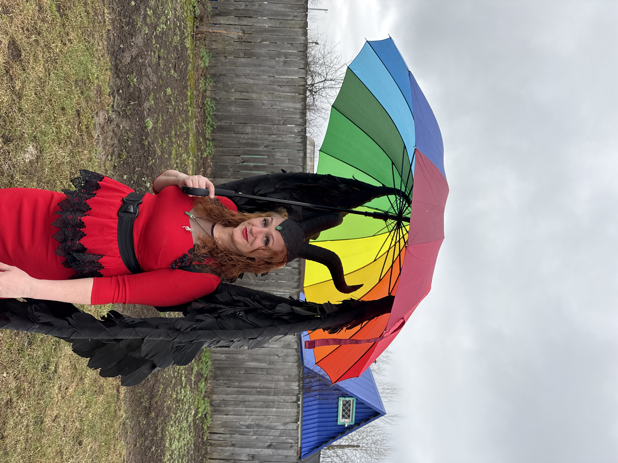 A woman stands outdoors holding a rainbow-colored umbrella. She is wearing a red dress with black lace trim, black wings, and horned headwear, suggesting a costume or cosplay. The backdrop includes a wooden fence, grass, and a small structure with a blue roof. The setting suggests an overcast day. Based on the reference to Chaikovsky, Russia, the picture was likely taken in or around that city.
