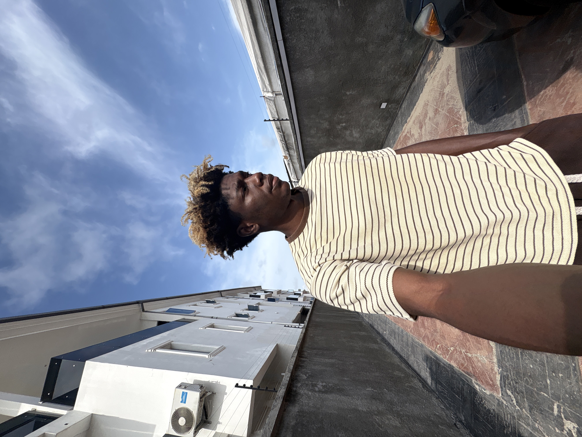 A low-angle shot depicts a man in a vertically-striped shirt standing outdoors in Igboefon, Nigeria. He has dark skin and short, curly hair that is blonde at the tips. He is looking towards the left of the frame. Behind him is a white, multi-story building with several windows and an air conditioning unit visible near the bottom. Above him is a blue sky with scattered white clouds. To his right, there is a concrete wall and a portion of a black car is visible. The ground appears to be paved with a mix of red and gray concrete or stone.