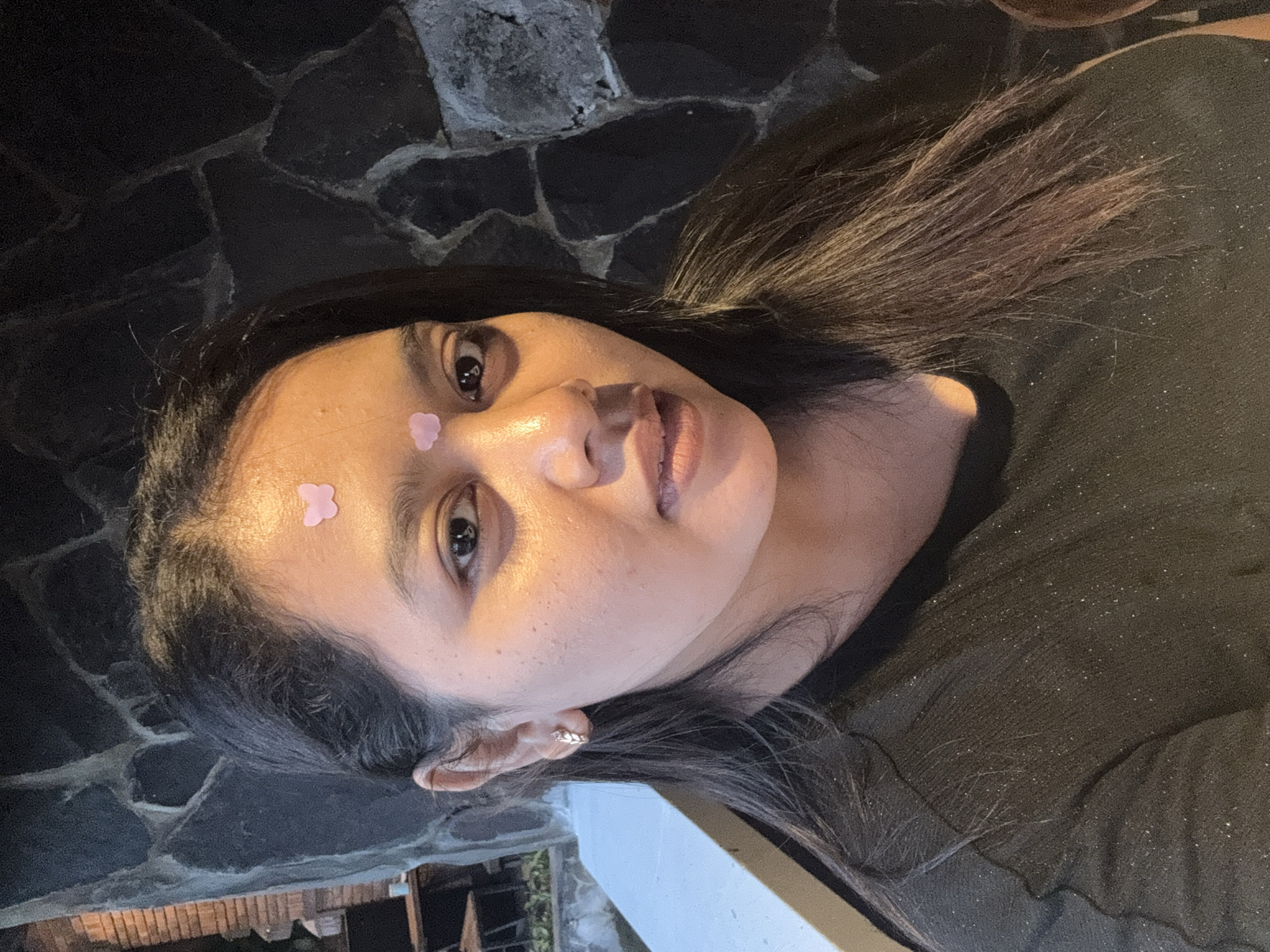 A woman with medium-length dark hair is positioned in the foreground, partially against a dark, roughly-textured wall. Two small, pink, flower-shaped stickers are adhered to her forehead. She is wearing a dark, possibly green, textured top. The lower part of the image shows portions of what may be wooden architectural elements, seen through what appears to be an outdoor setting. The location is inferred to be in Kuta Selatan, Indonesia.