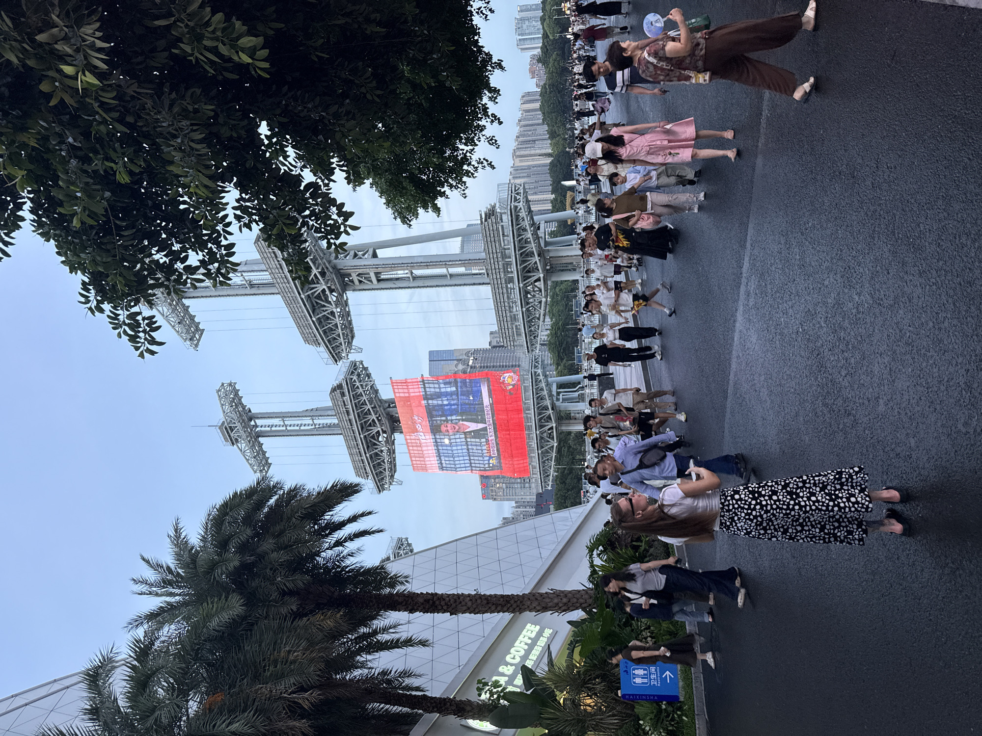 In Tianhe District, China, a public outdoor area features a large LED screen displaying a person's face. The screen is suspended between two large, industrial-style metal structures. A crowd of diverse individuals, including men, women, and children, are walking on a paved pathway in front of the screen and structures. Palm trees and leafy trees are visible on the left and right sides of the image, respectively. A building with a white, triangular facade is on the far left, with signage that appears to say "COFFEE." The sky is overcast and a pale blue. The ground is wet, indicating recent rain.