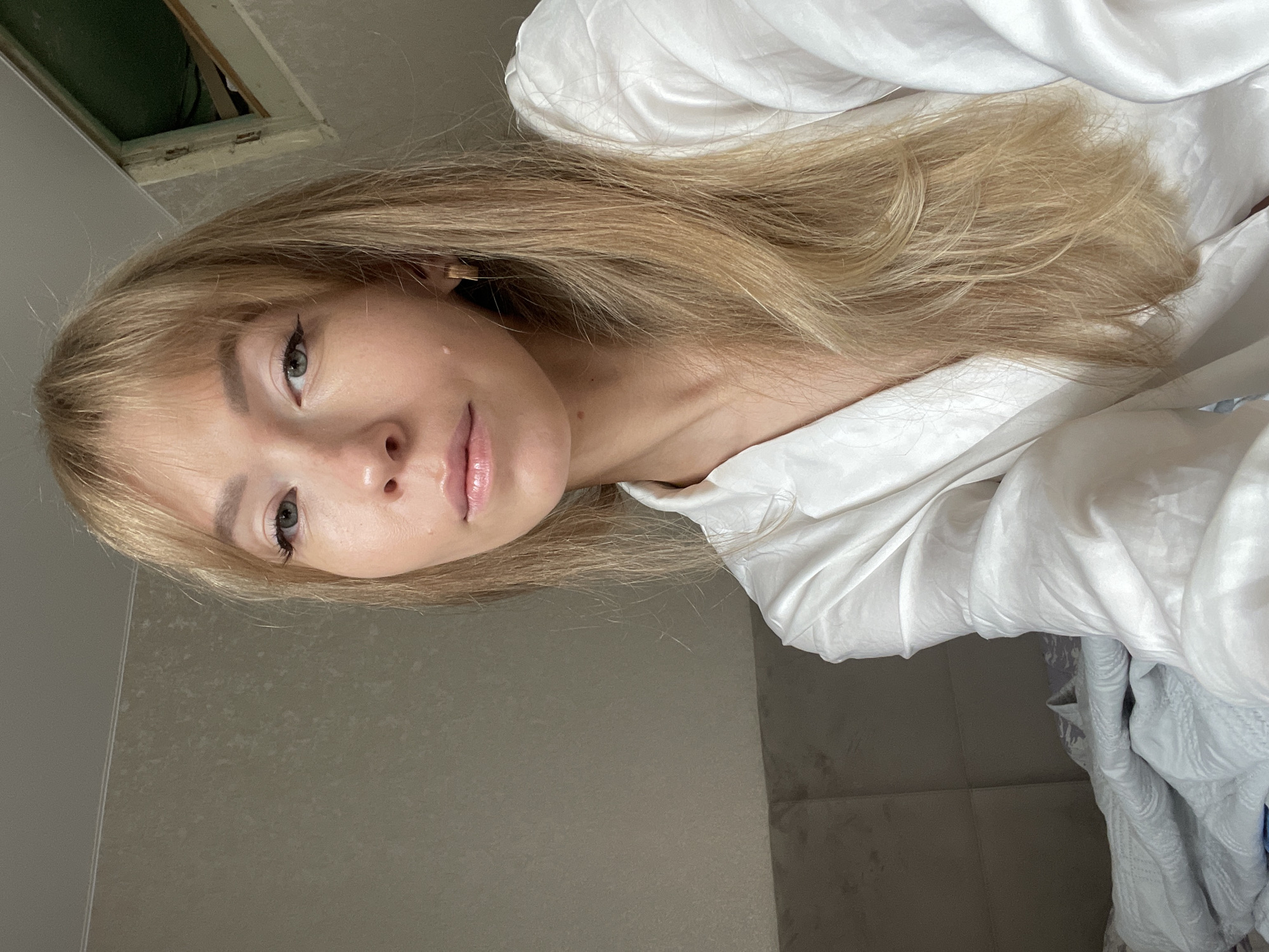 A young woman with long blonde hair and bangs, wearing a white silk robe, is in bed. Her hair is parted to the side and falls around her shoulders. She has light makeup on, including eyeliner. A small mole is visible on her left cheek, and a delicate gold earring is worn in her left ear. The background shows a wall with a textured pattern and a slightly damaged ceiling area. A portion of a headboard and white bedding are also visible. The setting appears to be an indoor bedroom, possibly in an apartment or hotel room. The lighting is soft and natural.