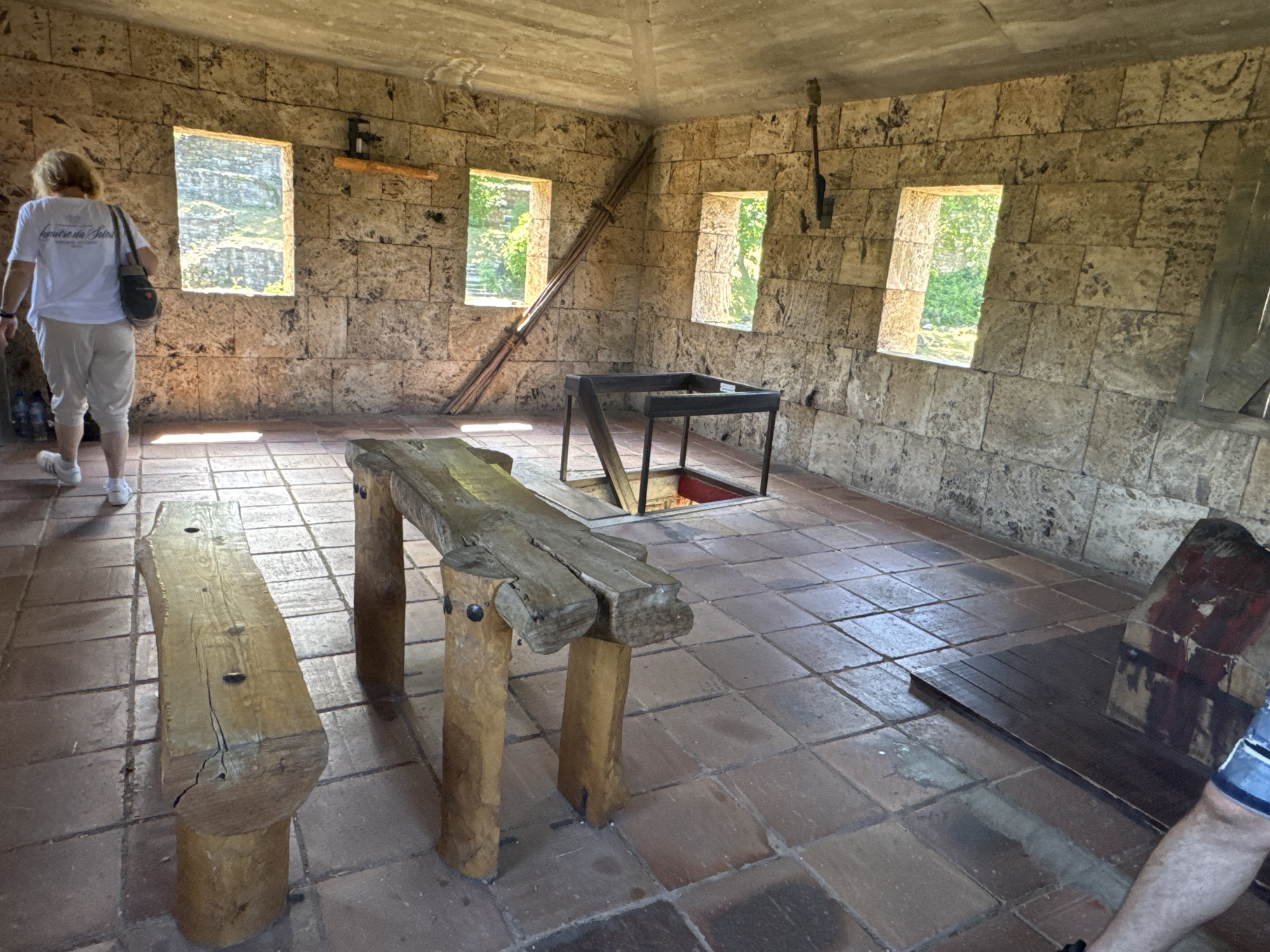 The image depicts the interior of a stone building with a rough-hewn stone block wall construction. The floor is tiled with square terracotta tiles. In the foreground, there is a rustic wooden bench with a rough-hewn surface and log-like legs. To the right of this bench is a longer, similar wooden bench or table. In the center of the room, a metal table with a square frame sits over a dark opening in the floor. Two rectangular windows with stone frames are visible on the left and right walls, letting in natural light. The window on the left looks out onto a rocky, sunlit landscape, while the window on the right shows greenery. A woman wearing a white t-shirt and light-colored pants, with a shoulder bag, is walking away from the camera on the left side of the frame. There are some tools or implements hanging on the wall near the ceiling. The overall impression is of a historical or rustic setting. This scene is likely located within the Tsarevets Fortress in Veliko Tarnovo, Bulgaria, as these types of rustic reconstructions of historical living or working spaces are common within the fortress grounds.