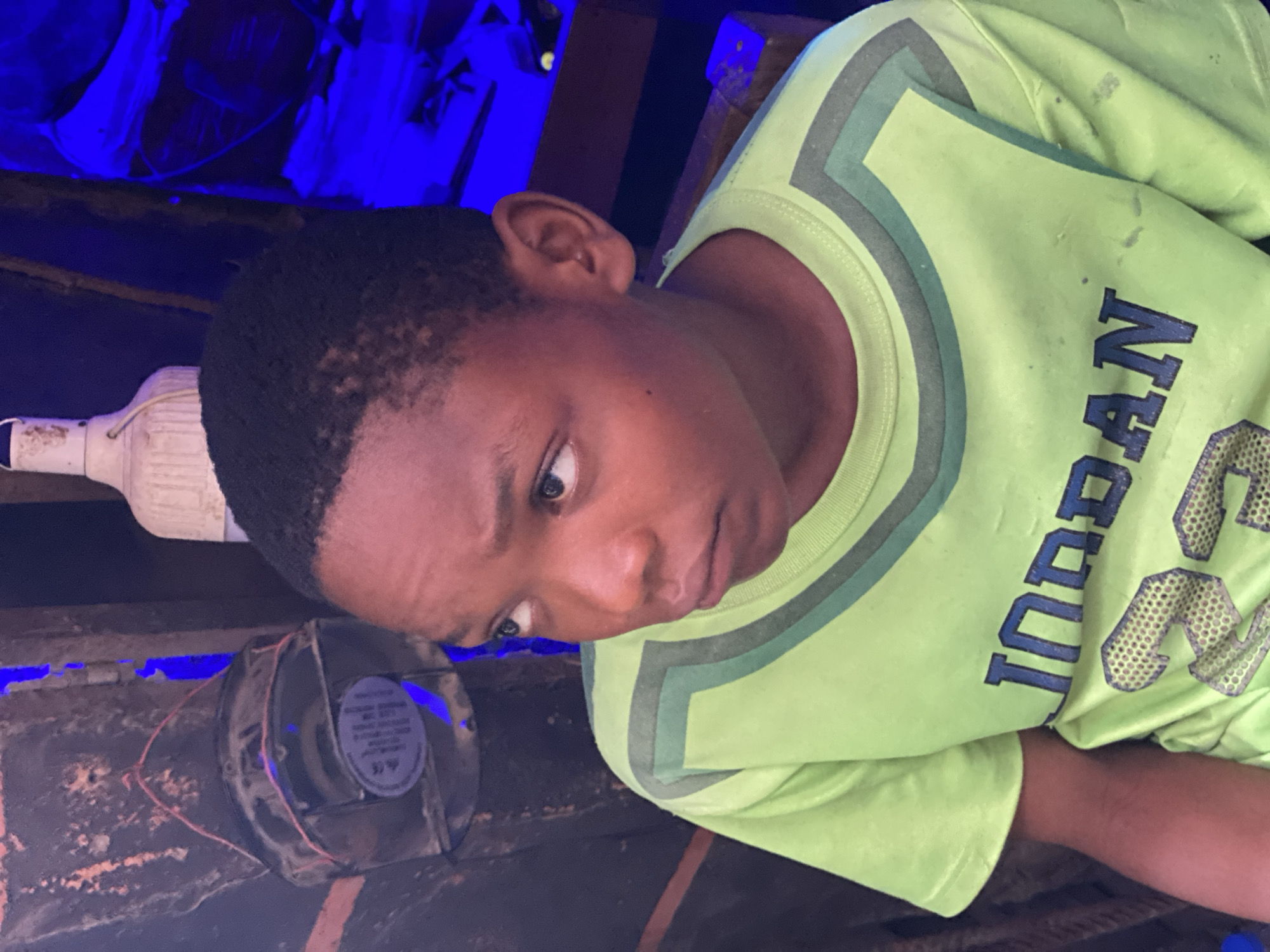 A close-up shot of a young Black boy with short, dark hair. He is wearing a light green t-shirt with "JORDAN" printed in blue letters and a number 23 made of mesh. The background features a dark wooden surface with a small fan attached and a white plastic bottle nearby. The lighting in the background is blue, creating an artificial ambiance. The location is possibly Fegge, Nigeria.