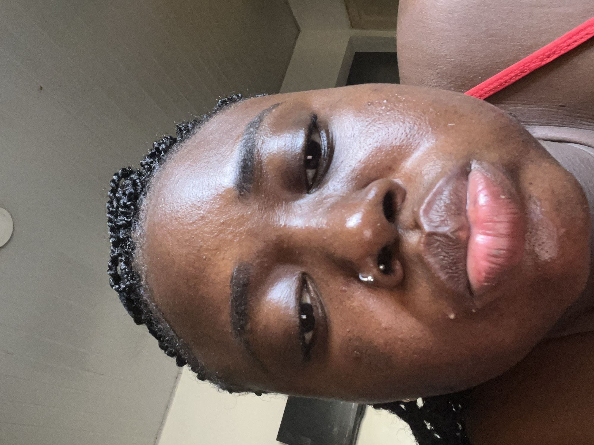 The image is a close-up, low-angle selfie of a Black woman with dark skin and braided hair. She is wearing a red top. Her face is tilted downwards towards the camera, and she is puckering her lips. The background appears to be an indoor setting with a textured ceiling and a wall. There is a dark rectangular object, possibly a television, visible to the left of the woman. There is no indication of the location being in Abuja, Nigeria; this information is not present in the image itself.